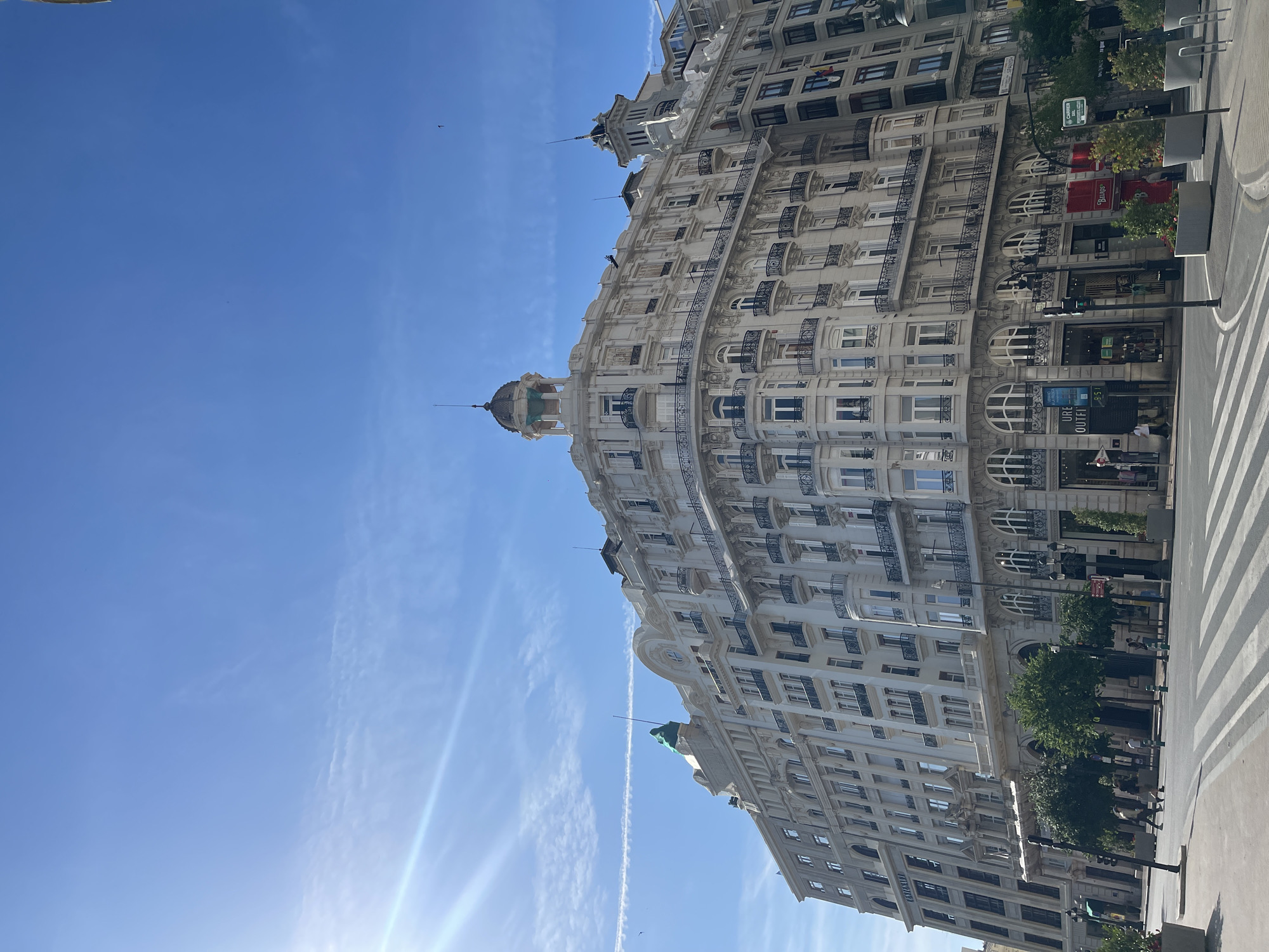 The image captures a grand, ornate building under a clear blue sky with subtle wispy clouds and faint contrails. The sun, positioned in the upper left quadrant, emits a visible lens flare. The scene is located in Valencia, Spain, specifically depicting the **Edificio Generali** (formerly Banco de Valencia) at the intersection of Carrer de Ribera and Carrer del Marquès de Sotelo.
The multi-story building is characterized by its light cream or off-white facade, featuring intricate architectural detailing including numerous balconies with black wrought-iron railings. The building's prominent curved corner is topped with a distinct dome-shaped structure, capped with a green or turquoise material and a pointed finial. Further right, another smaller, grey-domed turret is visible. The lower floors display arched windows, while upper levels have rectangular windows. The ground floor accommodates various commercial establishments, with "Urban Outfitters" and "BELROS" signage clearly visible. A small flag, consistent with the Valencian Community flag (red and yellow stripes with a blue stripe and crown), is visible hanging from an adjacent building on the far right.
In the foreground, a marked pedestrian crosswalk spans the street. Several streetlights, a traffic signal, and dark green bollards are positioned along the curb. Large grey planters containing small trees and greenery line the sidewalks. Approximately five to seven individuals are present, walking on the sidewalks or near the crosswalks. The overall impression is a vibrant urban street scene during daylight hours.