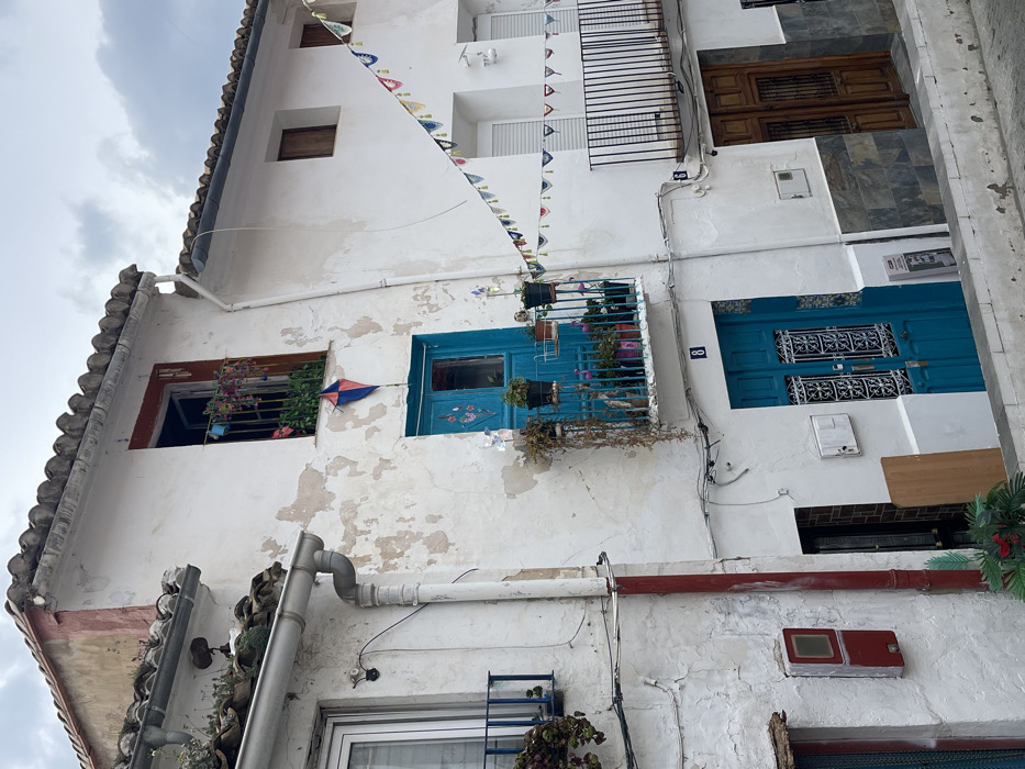 The image depicts a narrow street scene in Buñol, Spain, featuring three multi-story buildings under an overcast sky.
The central building, bearing the number '8' below its middle-floor balcony, has a weathered white facade with peeling paint and a terracotta tiled roof. The top floor features an open window with a red frame and a window box containing green foliage and several colorful pinwheel decorations. Directly below this window, a small red and blue diamond-shaped kite is affixed to the wall. The middle floor has a teal-blue double door with decorative grille work, opening onto a small balcony with a black metal railing. This balcony holds numerous potted plants, some with pink blooms, and two white pinwheel decorations. A white vertical drainpipe runs down the left side of this building, turning red at its lower section. The ground floor presents another teal-blue double door with similar decorative grille work. To the left of this door, a light-colored wooden board leans against the wall, and a red electrical box is mounted further left.
To the right, a multi-story building marked with the number '6' on its ground floor. Its upper section has a white facade, transitioning to dark grey, slate-like tiles on the lower half. The top floor features two small, dark-framed windows. The middle floor has two windows with closed white shutters and a balcony with a black metal railing, devoid of plants. The ground floor has a brown wooden double door with a small grille. A string of colorful, crescent-shaped fabric bunting hangs diagonally across the street, connecting the central building to this building on the right.
To the far left, a partially visible white building features a window with a blue metal balcony containing potted plants. No people are visible in the scene. The street surface is partially visible at the bottom, suggesting a curb or low wall in the foreground.