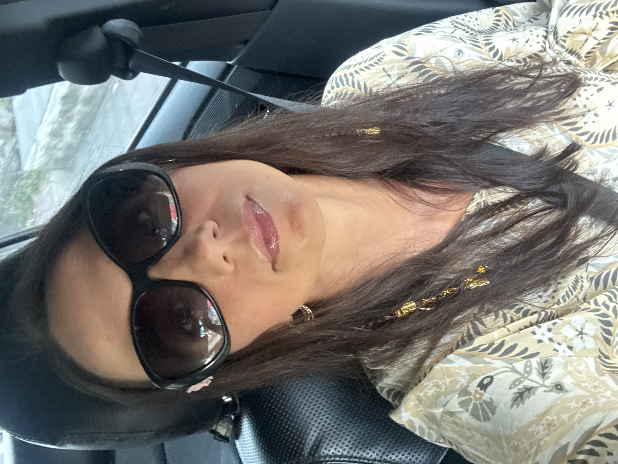 A woman wearing dark sunglasses and a patterned beige and gray top is seated in a car. She is wearing a seatbelt. Her location in Kuta Selatan, Indonesia is not determinable from the visual information.