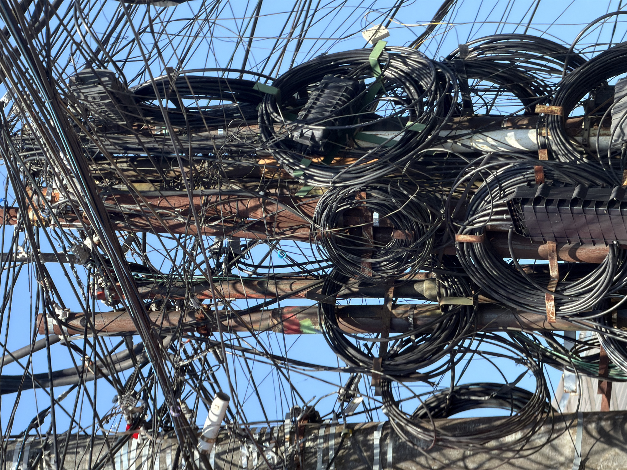 The image is a close-up, low-angle shot of a utility pole in Kuta Selatan, Indonesia, heavily burdened with a chaotic tangle of electrical and telecommunications cables. The pole itself appears to be made of metal, with some sections exhibiting rust and discoloration from weathering. Numerous black cables are coiled, bundled, and strung haphazardly across and around the pole. Some cables are thick and appear to be power lines, while others are thinner, possibly for internet or telephone service. Several cable management boxes, typically black and rectangular, are visible, some attached to the pole and others integrated into bundles of wires. There are also some metallic elements, like brackets and clamps, holding the cables in place. The background is a clear, bright blue sky, suggesting it is daytime. The overall impression is one of extreme disorganization and a dense, overwhelming network of infrastructure.