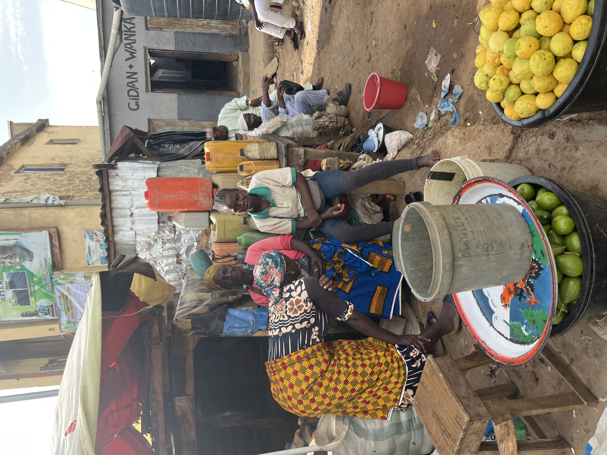 The image depicts an outdoor market scene in Rafin Par, Nigeria. In the foreground, a large black bucket is filled with yellow-green citrus fruits, likely lemons or limes, and another bucket contains similar green fruits. A red bucket is also visible on the ground. To the left, a small wooden stool sits next to a collection of large plastic containers, some red and some yellow, stacked near a stall with a tattered fabric roof.
Three women are the primary subjects in the center of the frame. One woman, wearing a pink shirt and a blue and patterned wrapper, is seated, holding a baby in her lap. Another woman, dressed in a blue and patterned wrapper, is bent over, seemingly tending to something or interacting with the seated woman. A third woman, in a white and green collared shirt and blue jeans, sits to the left, looking towards the viewer.
Behind the stall, several men are present. One man in a light green robe and a white cap is seated, gesturing with his hand. Another man in a white outfit and a cap is seated further back. The background shows buildings, one with the inscription "Gidan Wanka" above a doorway. The ground is dirt and there are various scattered items, including plastic bags and debris. The overall impression is a bustling, informal marketplace environment.