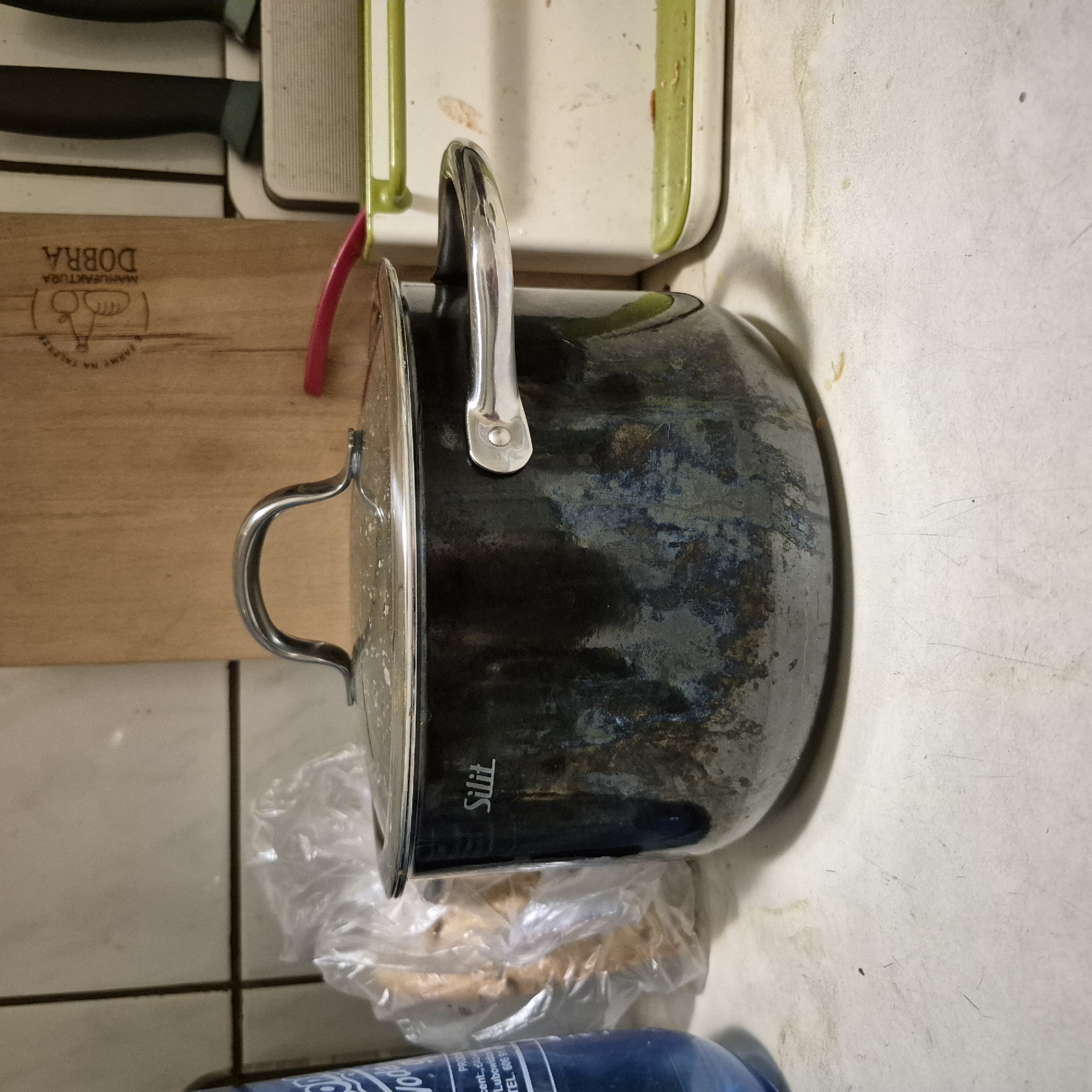 The image shows a dark, slightly discolored metal pot with a glass lid and a chrome handle, situated on a white countertop. The pot has the brand name "Silit" visible on its side. To the left of the pot, a blue plastic bottle with white lettering is partially visible, and behind it, a crumpled plastic bag containing some items. To the right of the pot, a white and green plastic container is present. In the background, a wooden cutting board with some markings and a red object are visible, along with a tiled wall. The scene appears to be a kitchen counter. There is no indication of the city of Kępno, Poland in the image or its context.