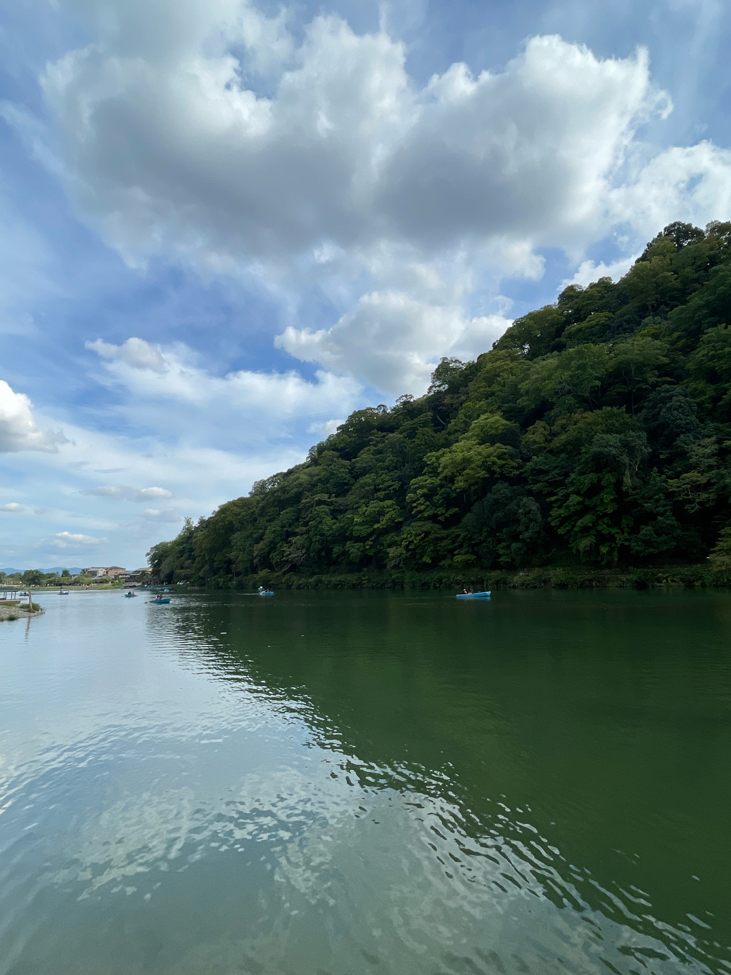 A scenic view of the Arashiyama Bamboo Grove area in Kyoto, Japan, features a tranquil river with several small boats on its surface. The river, with its calm, green water, reflects the overcast sky and the lush, verdant hillside that rises steeply on the right side of the frame. The hillside is densely covered in trees, creating a rich tapestry of green. The sky above is partly cloudy, with large, white clouds interspersed with patches of blue. In the distance, on the left bank of the river, a few buildings and trees are visible, suggesting a town or village. The overall impression is one of natural beauty and peaceful recreation.