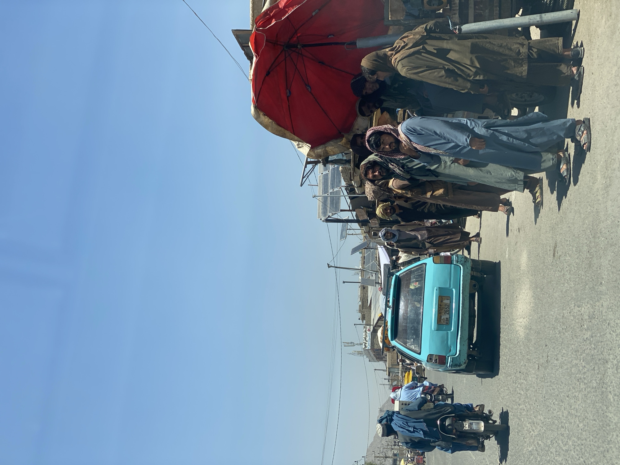 In Kandahar, Afghanistan, under a clear blue sky, a street scene unfolds with a mix of vehicles and pedestrians. A turquoise station wagon, with license plate KBR 2652, is positioned in the foreground on a paved road. A motorcyclist, wearing a blue tunic and headdress, is on the left, moving away from the viewer.
A group of men, predominantly in traditional Afghan attire including turbans and long tunics, are gathered on the right side of the road, some standing under a large, weathered red umbrella. They appear to be observing or waiting. Solar panels are visible on some of the structures in the background, suggesting a mix of modern and traditional infrastructure. The overall environment appears to be a bustling urban or semi-urban area, typical of a market or busy thoroughfare in Kandahar.