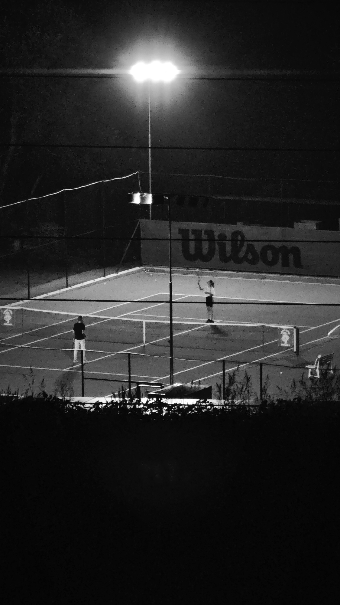 A black and white, medium-shot, eye-level photograph captures a tennis court at night, illuminated by a bright overhead lamp. Two individuals are visible on the court. On the left, a person in light-colored pants and a dark shirt stands facing the net. To the right, a person in dark shorts and a light top is in mid-serve, preparing to hit a tennis ball. The court itself is marked with white lines, and a net divides the playing area. In the background, a dark fence encloses the court, and a large "Wilson" logo is visible on a wall behind the fence. The foreground is dominated by a dark, silhouetted area of foliage. The overall setting appears to be an outdoor public space, potentially a park or sports facility, though no specific city or location details are discernible from the image itself.
