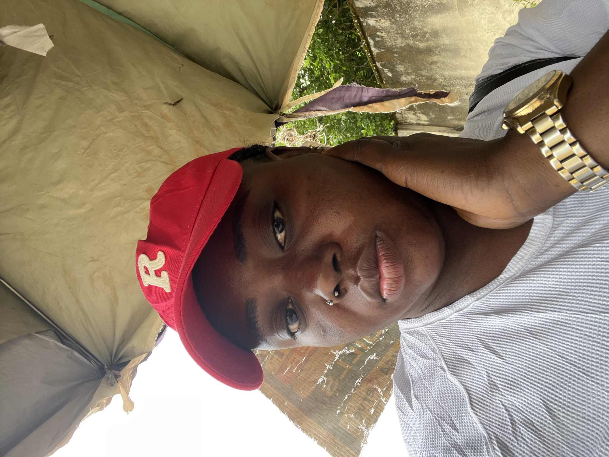 A close-up, eye-level, outdoor selfie shows a young Black person wearing a red baseball cap with a white "R" on the front. They are wearing a white waffle-knit shirt and a gold watch on their left wrist. Their left hand is touching their cheek. The background consists of a tan tarp overhead, a gray concrete wall, and green foliage. The image appears to be taken in Abuja, Nigeria, based on the context provided.