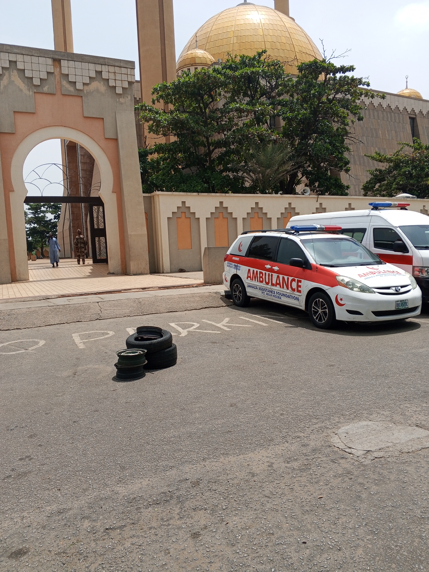 The image shows the National Mosque in Abuja, Nigeria. Two ambulances are parked next to the building, one a white minivan with red markings and "AMBULANCE" printed on the side, along with "JANAZA ASSISTANCE FOUNDATION, FCJ, ABUJA NIGERIA." The other ambulance is only partially visible. A stack of tires lies on the pavement in front of the ambulances, and two people are walking through the arched entrance of the mosque.