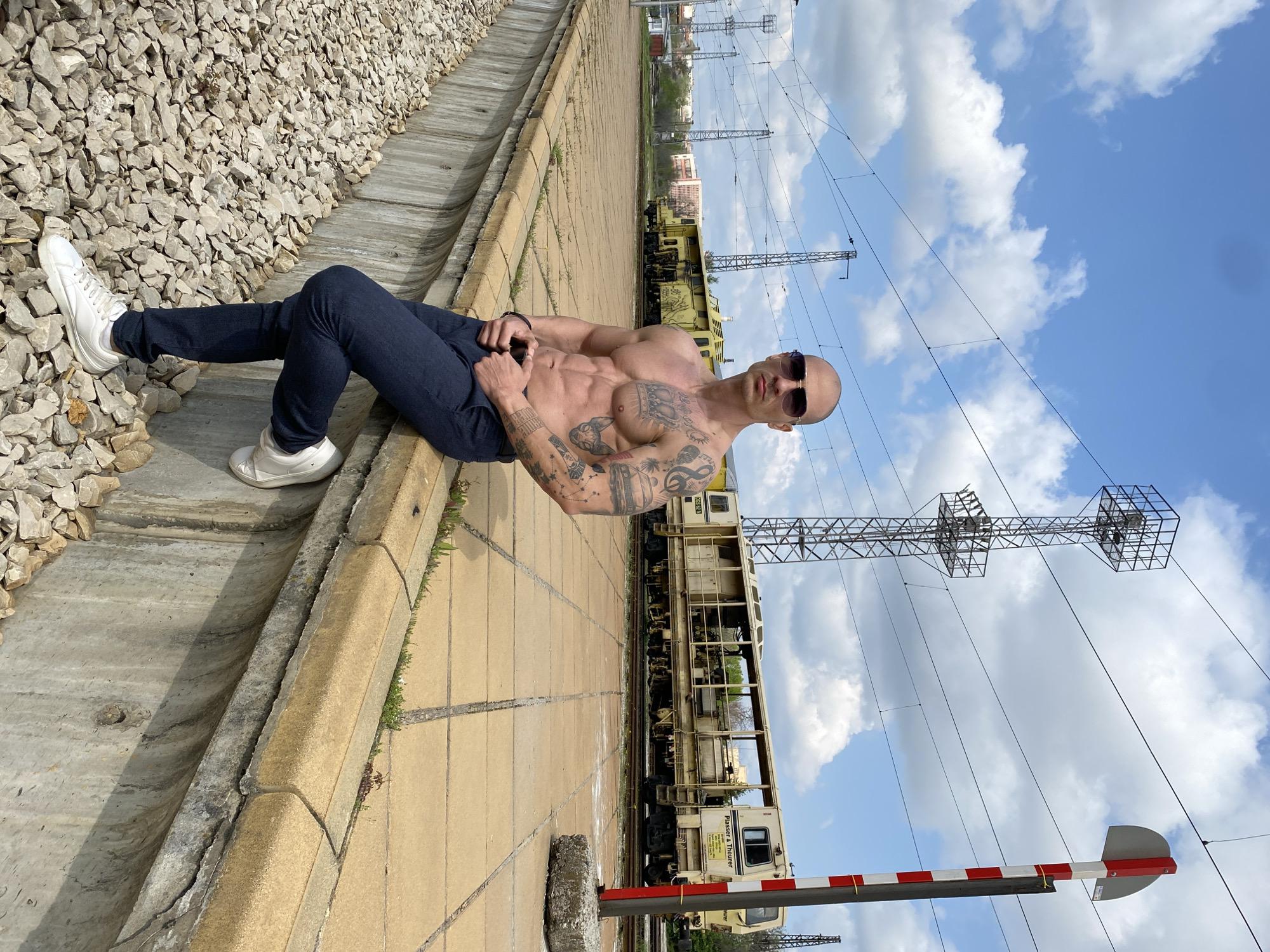 A muscular, tattooed man is seen reclining on a concrete surface next to a railway line in Pleven, Bulgaria, under a bright, partly cloudy sky. The man is shirtless, showcasing a well-defined physique and extensive tattoos covering his arms and torso. He wears dark blue trousers, white sneakers, and dark sunglasses, looking directly at the viewer with a neutral expression.
He is positioned with his back against a sloped embankment of light-colored gravel. Adjacent to him are train tracks, on which a long, yellow railway maintenance vehicle, identified as a "Plasser & Theurer" machine, is visible in the mid-ground. Overhead, a complex network of power lines and robust metal gantry structures crisscross the sky. In the lower right corner, a red and white striped railway barrier is partially visible. The scene is an outdoor, industrial setting, likely a train station or maintenance yard, during daytime with clear weather conditions. Visible text on the yellow train includes "0832" and "SYSTEM".