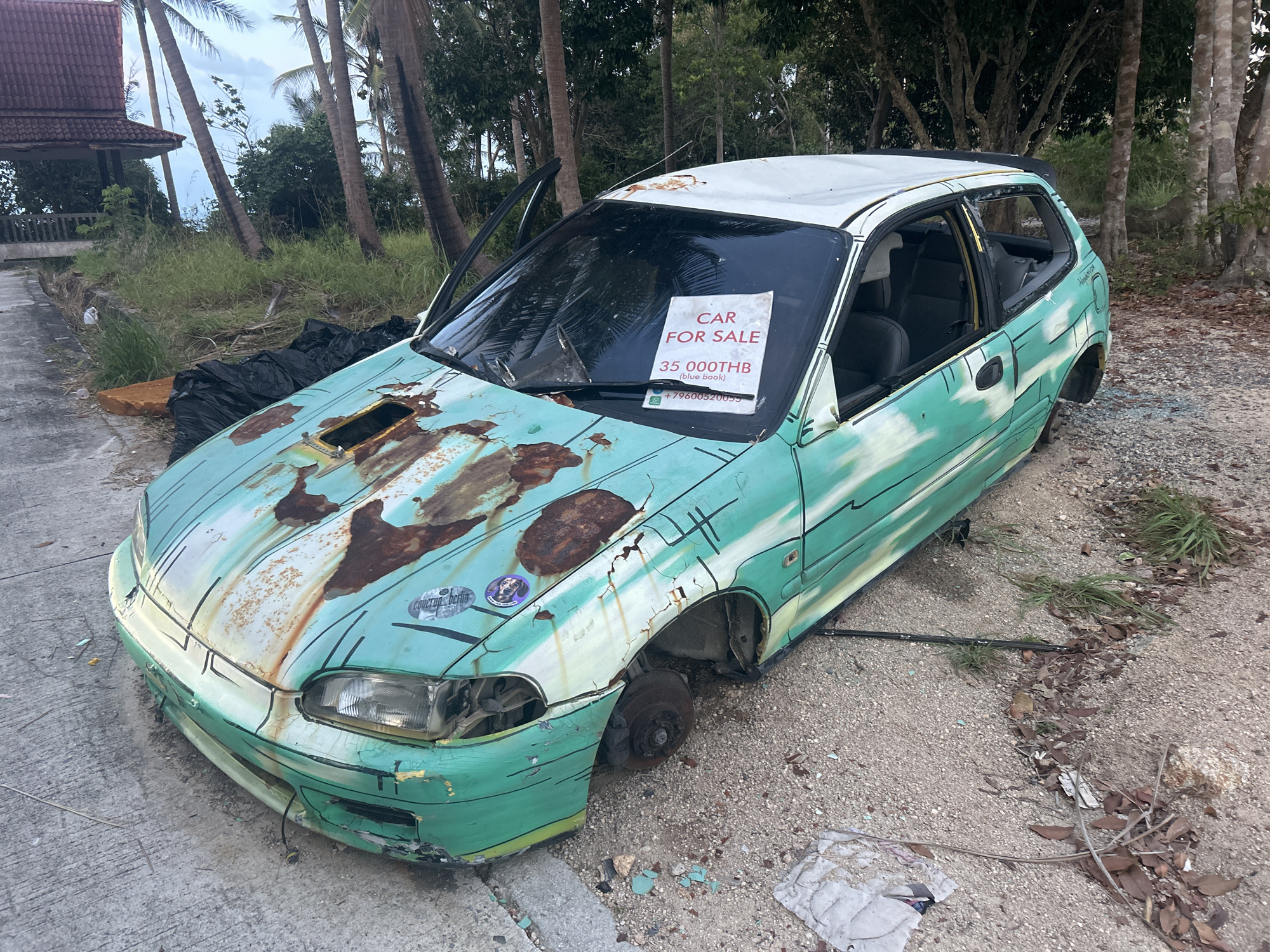 The image shows a dilapidated, light green and white, four-door hatchback car for sale, located outdoors. The car is heavily rusted, particularly on the hood, and missing wheels, exterior trim, and window glass. The driver's side door is open. A "CAR FOR SALE" sign, priced at 35,000 THB (Thai Baht), is displayed on the windshield. The car is situated on a gravelly or sandy surface with scattered debris and some vegetation. Background features include trees, a partial view of a building or structure, and the presence of grass. The scene appears to be in Baan Wang Ta Kien, Thailand.