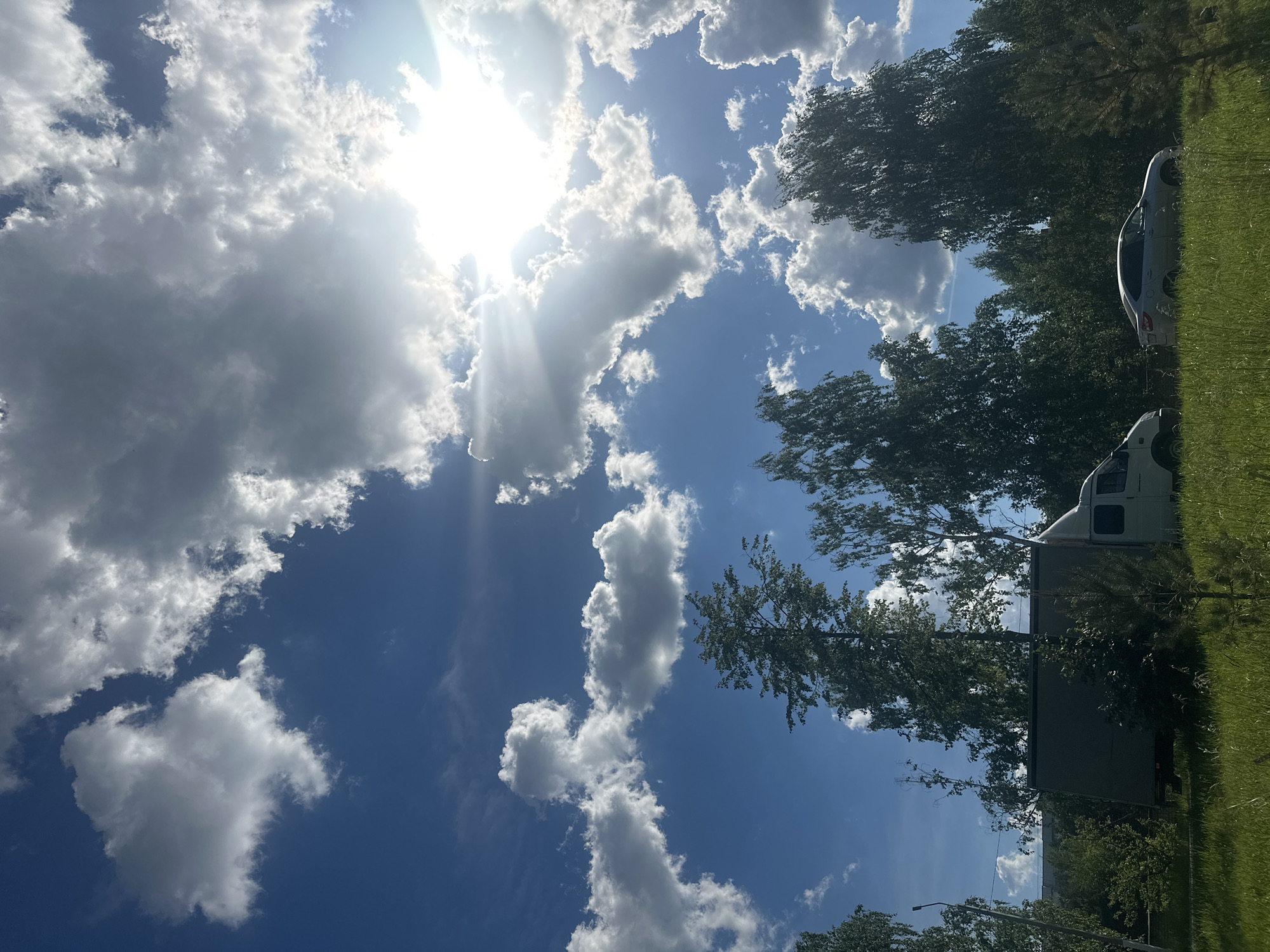 This is a vertical, medium-shot, outdoor photograph captured on a sunny day in Chaikovsky, Russia. The sky is predominantly blue with scattered white cumulus clouds. Sunlight streams from the upper right quadrant of the image, creating lens flare and highlighting some of the clouds. In the foreground, a patch of tall, green grass is visible. Behind the grass, a line of various trees with lush green foliage forms a dense line. A white delivery truck is parked on the left side of the image, facing right, with its side visible. To the right of the truck, a silver sedan is parked, also facing right. The overall impression is a sunny, natural environment with vehicles present, likely on the outskirts of the city.