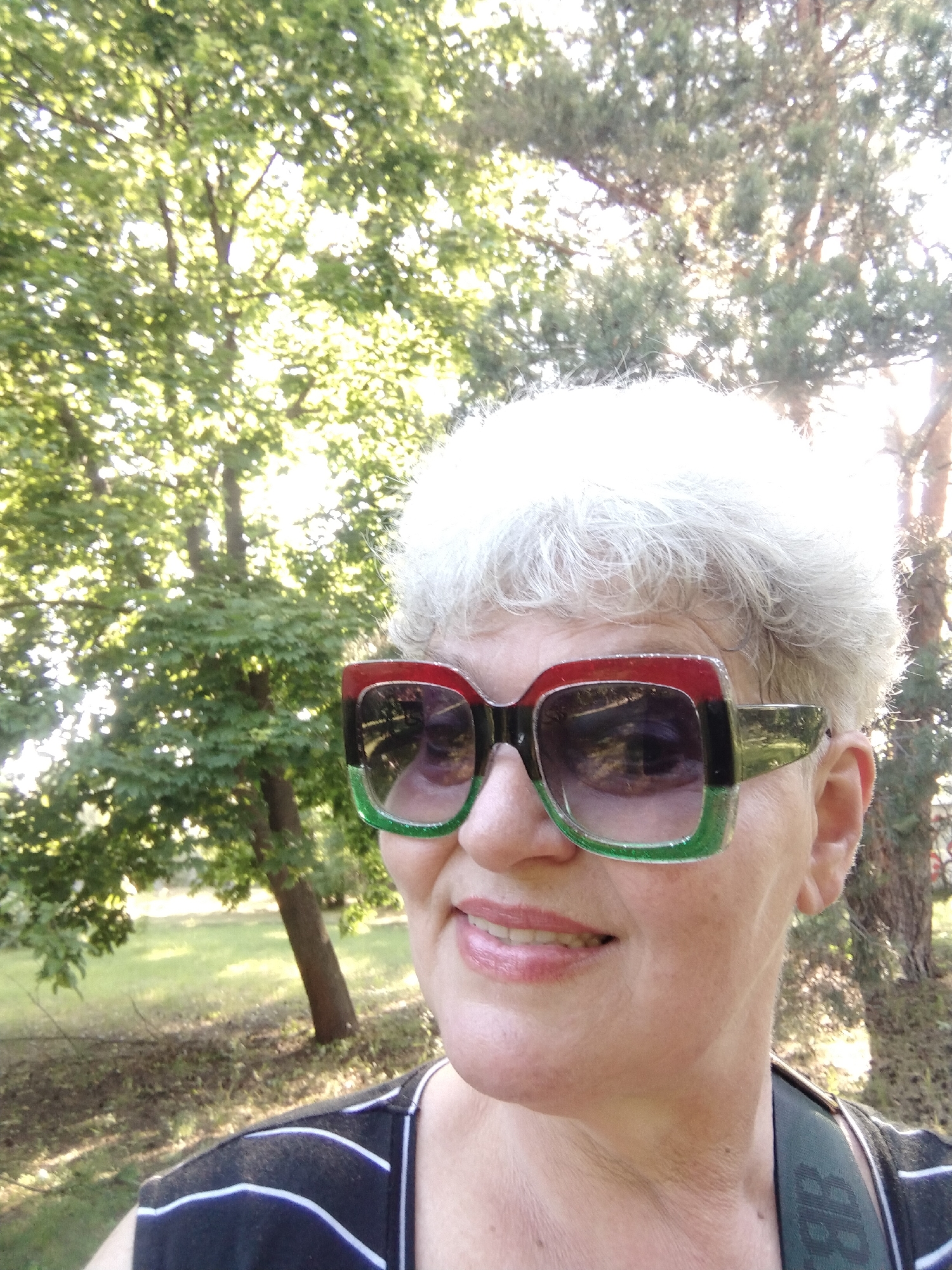 A fair-skinned individual with short, wavy, gray hair is depicted in a close-up outdoor portrait taken in Horishni Plavni, Ukraine. The person is smiling faintly, looking slightly upwards and to the right of the frame. They are wearing oversized, square-framed sunglasses with a distinct multi-colored frame: red on the top edge, black in the middle, and a glittery green on the bottom edge. The dark, reflective lenses show a blurred reflection of the surrounding trees and sky. The person's lower lip is visible, showing a pinkish gloss. They are dressed in a black top with white horizontal stripes across the chest area, and a black strap with faint white lettering is visible over the right shoulder. The background consists of lush green deciduous trees with sunlight filtering through their leaves on the left, and a darker green coniferous tree on the right, with strong backlighting from the sun causing significant glare and brightness. The ground visible behind the individual is grassy.