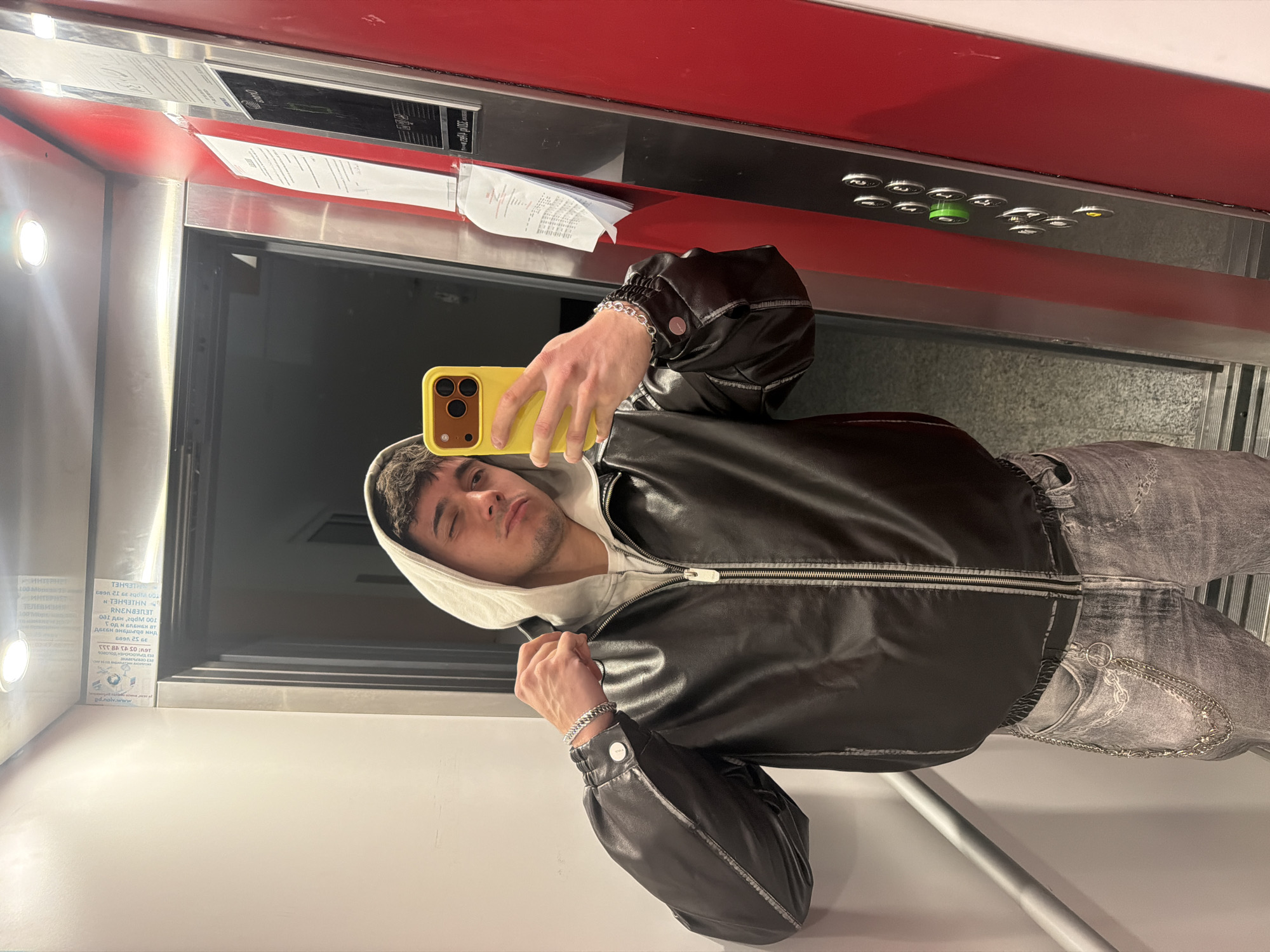 A young man is captured in a mirror selfie inside an elevator, the image rotated 90 degrees clockwise. He has short, dark, curly hair and appears to be winking or has one eye closed, with a neutral to slightly smirking expression. He wears a light-colored hooded sweatshirt under a black, possibly faux leather, zippered jacket. His attire includes grey, distressed jeans adorned with a silver chain. A silver chain bracelet is visible on his left wrist as he holds a yellow smartphone, featuring a prominent camera bump, to take the picture.
The setting is an elevator interior, featuring a bright red panel at the top, beneath which lies a dark control panel with numerous silver buttons, one illuminated green and another yellowish. Reflected in the mirror are various notices and papers. A bright light fixture is visible on the upper left side of the reflection. The bottom left shows a plain white wall panel.
Visible text, likely reversed from reflections, includes fragmented words such as "pd.nolv.www", "RENAT", "RENATEL", "NHIEbHELN", and "RIEBHEL", along with numerical sequences like "39 52 1609". These likely originate from emergency instructions or information placards within the elevator. The scene takes place indoors, illuminated by artificial light, in an elevator located in Sofia, Bulgaria.
