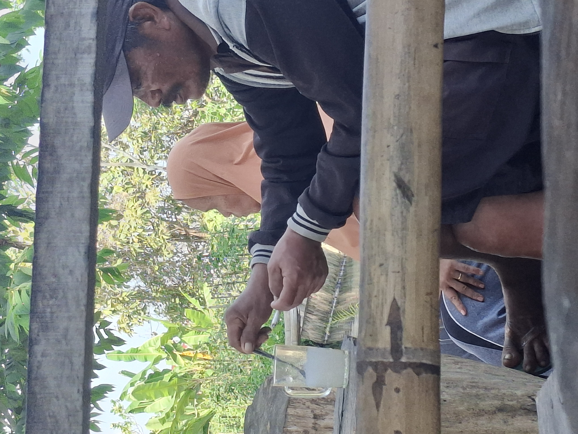 In Jogotrunan, Indonesia, two individuals are seated outdoors, possibly under a shaded structure constructed of rough-hewn wooden planks. In the foreground, a man wearing a baseball cap and a jacket with white and black striped cuffs is holding a cigarette in his right hand and a spoon in his left, stirring a white liquid in a clear glass mug. Beside him, a person wearing an orange headscarf is visible from the side. The background features lush green foliage, including banana leaves and other trees, under a bright sky. The lower portion of the image shows a person's bare foot and hand, the latter adorned with a ring, resting on gray fabric.