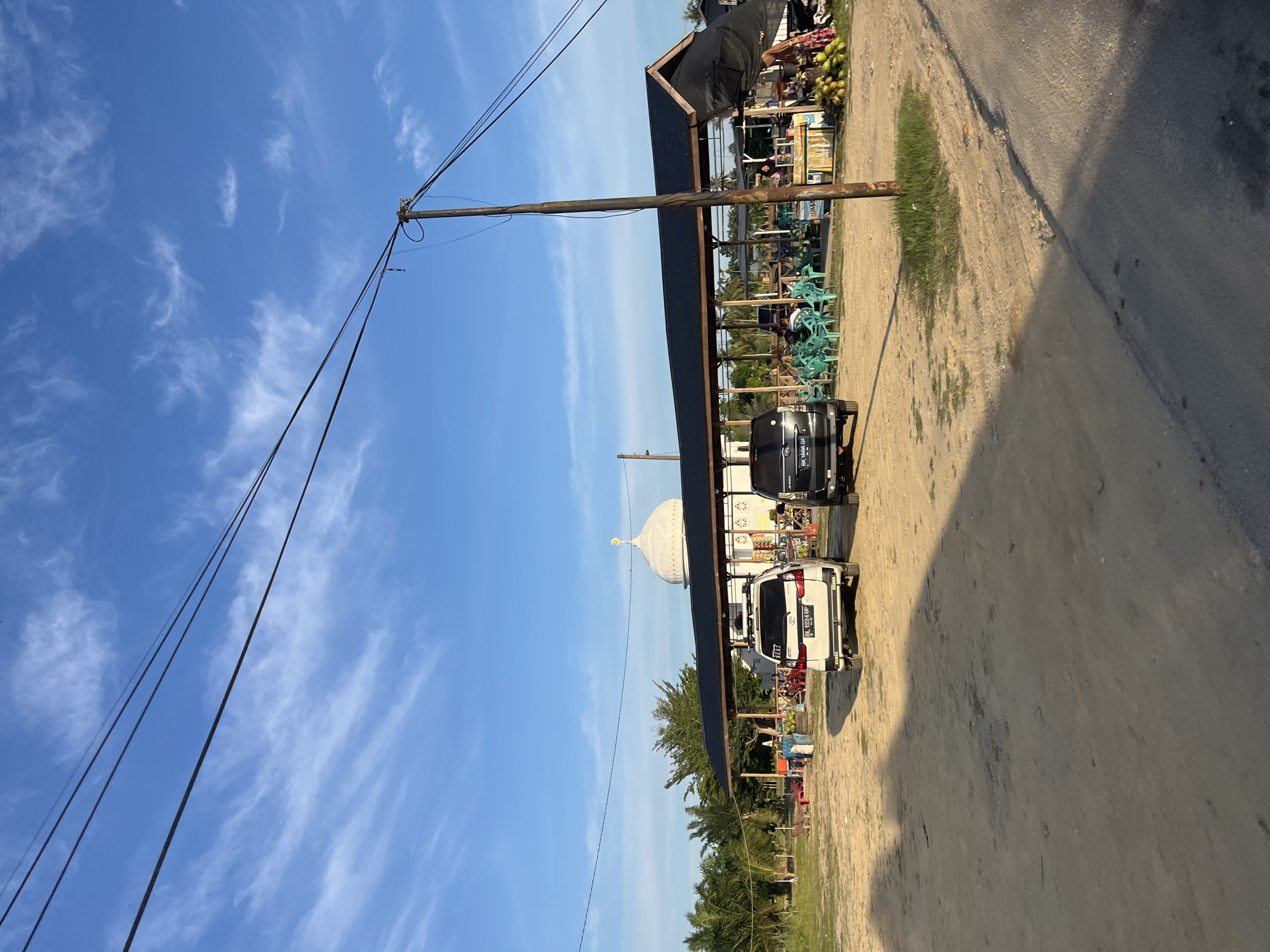 The image shows an outdoor scene on a bright, sunny day with a clear blue sky and scattered wispy clouds. The camera is positioned at an angle, giving a sense of looking up and to the side.
In the foreground, a wooden utility pole stands tall, with several dark electrical wires extending from it. The ground appears to be a mix of sandy soil and some patches of grass. A dirt road is visible on the right side of the image.
A long, dark-roofed structure, possibly a market or a building with an awning, stretches across the middle of the frame. Underneath this structure, several bright green plastic chairs are neatly arranged, suggesting a seating area or an outdoor dining space. A white SUV with a license plate reading "BK 2224 IIF" is parked near the structure. Further back, a black car is also visible.
Beyond the structure, a white dome with a golden finial, resembling a mosque or a similar religious building, is visible against the sky. In the background, there are trees and more distant structures. On the left side of the structure, there are stalls displaying what appear to be produce, possibly coconuts, and other goods. A few people are present, though their features are not clearly discernible.
The lighting suggests it is daytime, likely late afternoon given the long shadows. The overall atmosphere appears to be calm and perhaps a bit lively due to the market setting. The location context points to Seuneubokpunti, Indonesia.