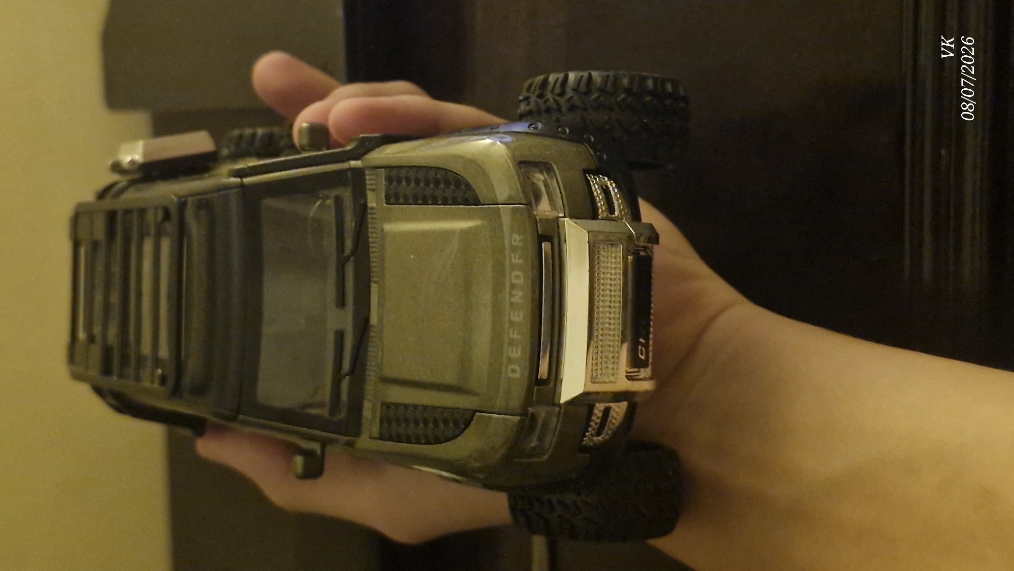 A hand holds a green toy SUV, possibly a model of a Land Rover Defender, with a roof rack. The toy has detailed features like a grille, headlights, and wheels. The background is blurred and appears to be indoors, with a wooden surface and a yellow wall. A watermark in the lower right corner reads "VK 08/07/2026". The image was taken in the city of Vrindavan, India.