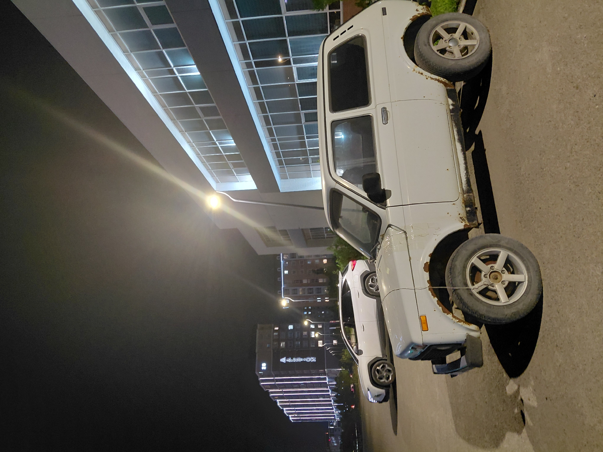 In the city of Aktau, Kazakhstan, at night, two cars are parked on an asphalt surface in front of a modern building with large windows. In the foreground, a white, older model SUV with visible rust on its body and wheel arches is parked. Its tires are dusty, and the vehicle appears weathered. Behind it, a white, modern SUV is parked, facing to the left. In the background, a tall, multi-story building is illuminated by strings of white lights that form vertical lines. The building has the word "ALBION" displayed in large, illuminated letters. The sky is dark, suggesting it is nighttime. Streetlights cast light on the scene.
