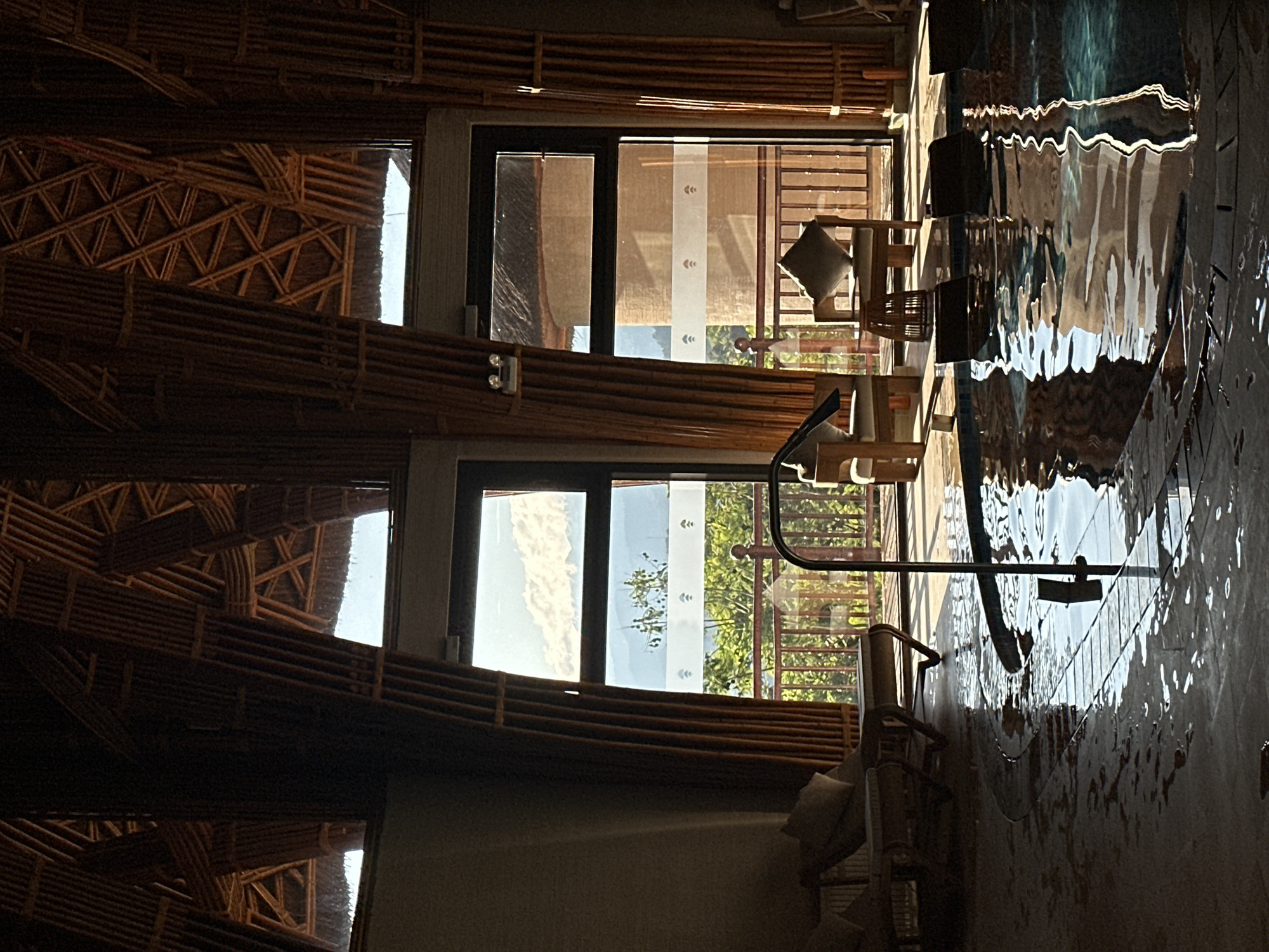 The image captures an inviting indoor space, likely within a resort or lodge in Mu Cang Chai, Vietnam. The scene is dominated by natural materials, with walls and ceilings constructed from intricately arranged bamboo or wooden slats, forming various geometric patterns. This rustic yet sophisticated design element creates a warm and earthy ambiance.
Large, dark-framed windows or glass doors open up to a bright outdoor view featuring lush green foliage and a clear, blue sky, indicative of a serene natural landscape. Through one of the windows, a blind with a distinctive white vertical strip adorned with small, repeating decorative motifs is visible.
In the foreground and extending to the right, a significant body of water, presumably an indoor or semi-indoor swimming pool, reflects the surrounding environment. The rippling surface beautifully distorts the reflections of the bamboo structures and the outdoor scenery into abstract patterns of light and shadow. A metal handrail or piece of equipment is positioned near or within the water.
Wooden furniture, including chairs with light-colored cushions, is arranged near the windows, suggesting comfortable seating areas for relaxation. No people are present, contributing to the tranquil and undisturbed atmosphere. The bright sunlight streaming through the windows and reflecting off the water indicates a daytime setting, likely mid-morning or afternoon, under clear and sunny weather conditions. The overall impression is one of peace, relaxation, and harmony with nature, consistent with a retreat in a scenic location like Mu Cang Chai. No discernible text is visible within the image.