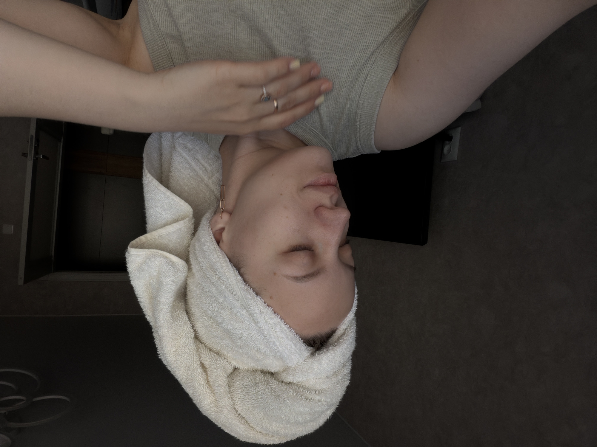 The image shows a person lying down with a white towel wrapped around their head. The person is wearing a light-colored shirt, and a hand with rings is touching their upper chest/neck area. The background is a dark, somewhat textured surface. The location in Reshetnikovo, Russia is not determinable from the image.