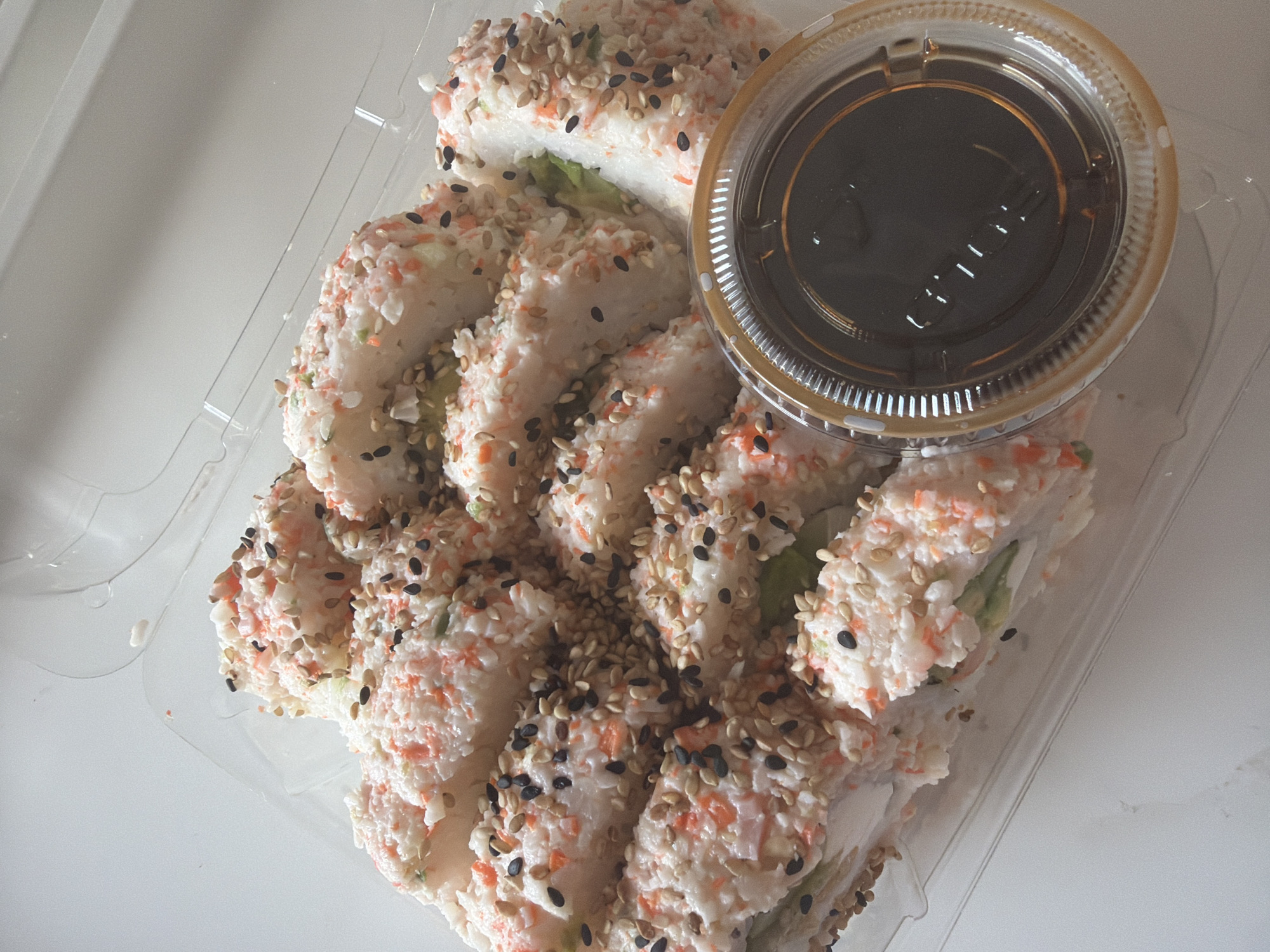 A high-angle, eye-level close-up shot shows a take-out container of sushi and soy sauce. The sushi appears to be California rolls, coated in imitation crab meat and sprinkled with black and tan sesame seeds. The rolls are cut into individual pieces and are arranged in a row, with some overlapping. To the right of the sushi is a clear plastic container filled with dark soy sauce. Both the sushi and soy sauce are contained in a clear plastic clamshell container. The background is a plain white surface. Location is unknown.