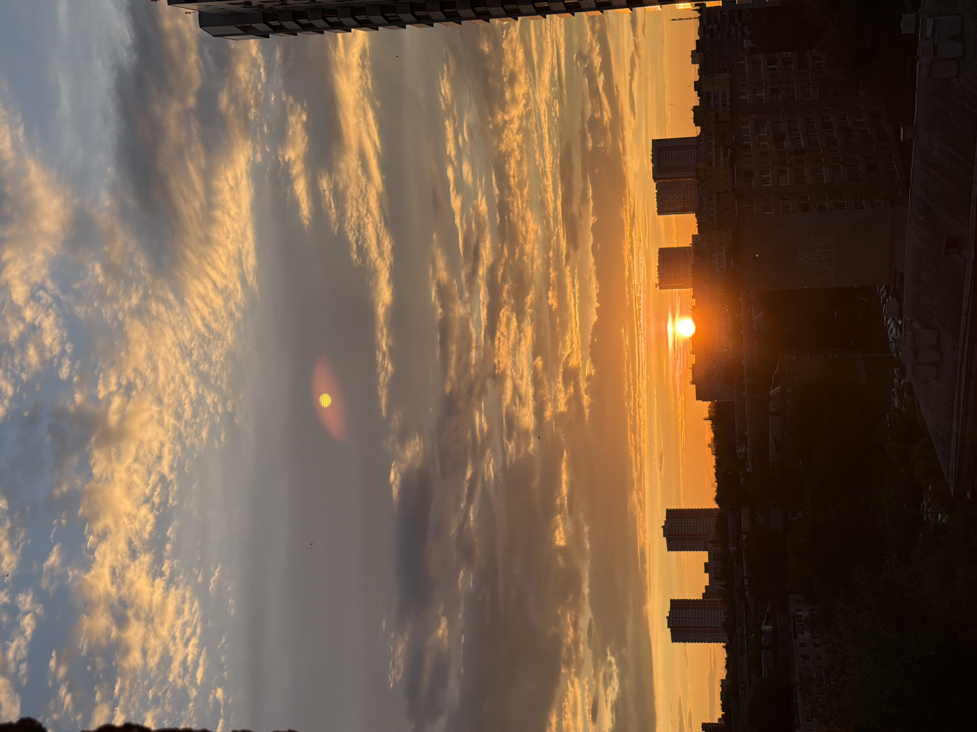 The image displays a dramatic urban landscape at either sunrise or sunset, characterized by a vibrant and cloud-filled sky. The horizon is punctuated by the silhouettes of numerous city buildings, including high-rise apartment blocks and other structures, rendered in deep shadow due to the strong backlighting.
A prominent, bright orange sun is visible low on the horizon, partially obscured by the dark outlines of the city. Its intense glow illuminates the surrounding clouds, painting them in striking shades of orange, yellow, and gold, creating a contrast with the cooler grey and light blue tones in other parts of the sky. The clouds exhibit varied formations, from wispy streaks to denser, more textured masses.
Higher in the sky, another distinct, circular yellowish light source is present. This object displays a noticeable grid-like pattern, suggesting it is an optical artifact such as a lens flare or internal reflection rather than a celestial body. Several tiny, dark specks, likely birds, are visible against the bright sky. The scene evokes the grandeur of an urban dawn or dusk, with no discernible human activity or visible text.