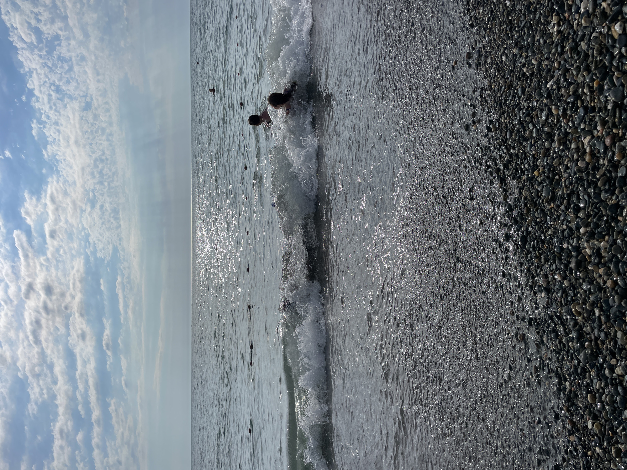 The image depicts a sunny day at a pebble beach in Sochi, Russia, with two individuals swimming in the moderate waves of the Black Sea. The water sparkles under the sunlight, reflecting the blue sky dotted with scattered clouds. The foreground is dominated by a dense layer of dark, smooth pebbles that transition into the water. A rope with buoys stretches across the water in the middle ground, indicating a designated swimming area. In the distance, a few other swimmers are visible, and the horizon line separates the sea from the sky. The overall scene is one of recreational activity in a natural coastal environment.