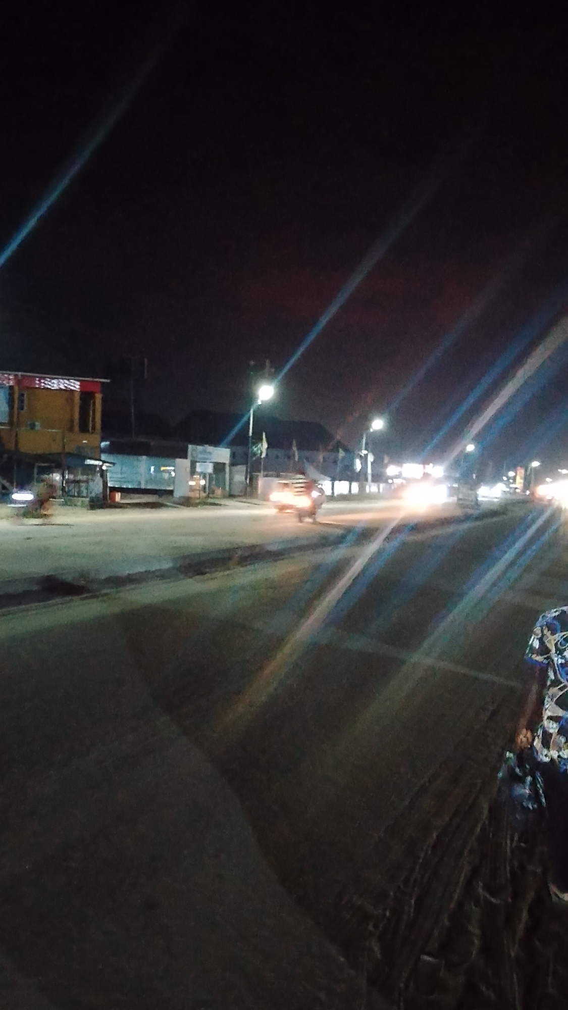 The image shows a street scene at night in Ohoro, Nigeria. Streetlights illuminate the road and surrounding buildings. There is a yellow building on the left side of the image. Traffic is present, with a vehicle in the middle ground. A person is visible in the lower right corner.