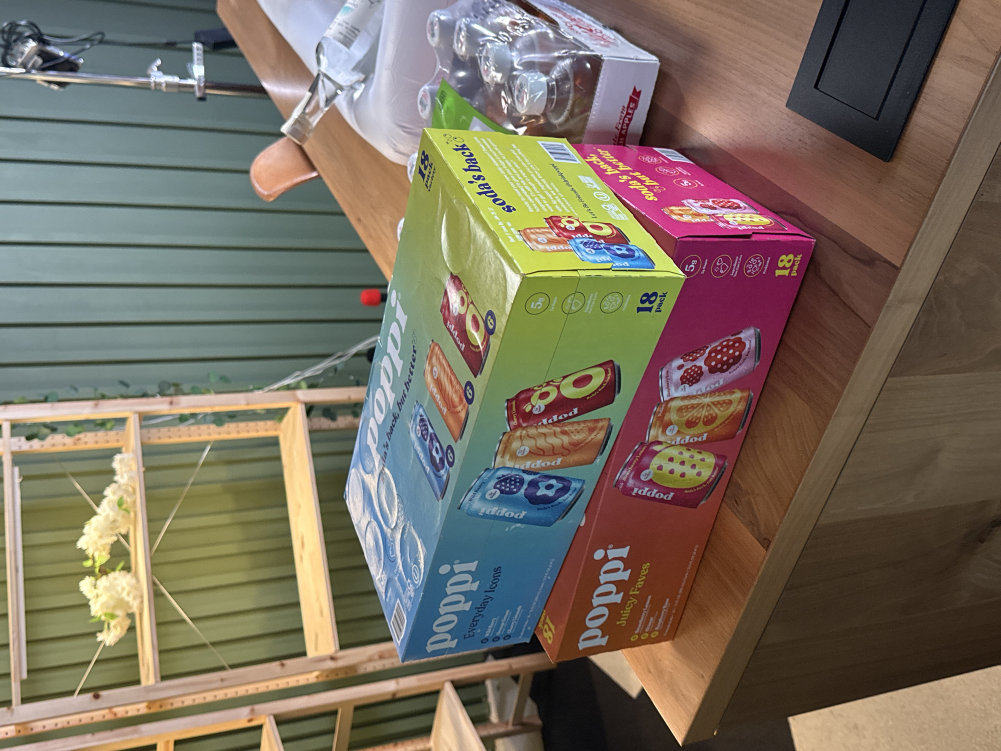 Here is a detailed description of the image:
**General Description:**
The image shows a close-up of a wooden table surface on which two boxes of "Poppi" sparkling prebiotic soda are placed. The boxes are situated next to a pack of glass bottles, seemingly containing a similar beverage. The background includes a wall with horizontal panels and a wooden structure with some floral decorations.
**Detailed Analysis:**
* **Foreground:**
* Two boxes of "Poppi" soda are the primary focus. One box, predominantly blue and green, is labeled "Everyday Icons." The other box, pink and orange, is labeled "Juicy Faves." Both boxes indicate that they contain 18 cans of soda.
* Behind the Poppi boxes, there is a clear plastic package containing glass bottles.
* A small black rectangle, possibly a coaster or a decorative element, is visible on the upper right corner of the table.
* **Background:**
* The wall in the background is made of wooden panels arranged horizontally. The panels appear to be painted in a muted green or grayish-green color.
* A wooden structure, possibly a trellis or part of a decorative installation, is partially visible. It has a grid-like design.
* White floral decorations are attached to the wooden structure.
* **Environment and Lighting:**
* The scene appears to be indoors, possibly in a studio or a staged area.
* The lighting seems to be soft and diffused, with no strong shadows.
* **Additional details:**
* There are some lighting equipment in the top left corner.
* It is evident that the scene has been rotated, as the horizontal wall panels appear to be vertical.
**Location:**
Given the presence of Poppi soda, a product commonly available in the United States, the scene likely takes place in the USA. Without more details, the exact location cannot be determined solely from the image.