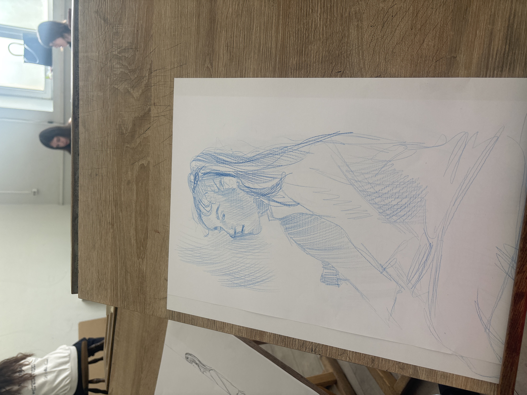 The image captures an indoor scene, likely an art studio or classroom, illuminated by natural light from a window. In the foreground, a light-toned wooden table or desk fills the right side of the frame. On this surface, a sheet of white paper displays a blue pencil sketch of a person, possibly a woman, with long hair, depicted in a pensive or relaxed pose with her head tilted downwards. The drawing utilizes loose, gestural lines and hatching for shading, conveying a sense of melancholy or introspection.
Reflected in the window, visible on the left side of the wooden surface, are the partial figures of two individuals. One reflection shows a head and shoulders, while a second, lower reflection reveals just a head. In the background, further to the left, a person with long, dark hair is seen from behind, wearing a white t-shirt with some unreadable text. This individual appears to be sitting on a chair or bending over, possibly engaged in an artistic activity or observation. A portion of another sketch, depicting a human figure, is visible on the table beneath the main drawing. The overall scene conveys a calm, creative atmosphere focused on artistic practice.