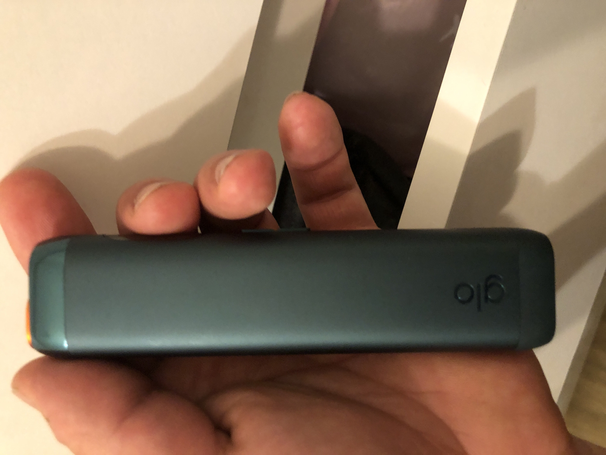 A person's hand is holding a teal-colored electronic smoking device with the logo "glo" embossed on its side. The device has a bright orange tip. The background appears to be a white box or surface, with a portion of the device partially visible within it. The image is a close-up shot, focusing on the device and the hand. No specific location in Leszno, Poland can be determined from the image.