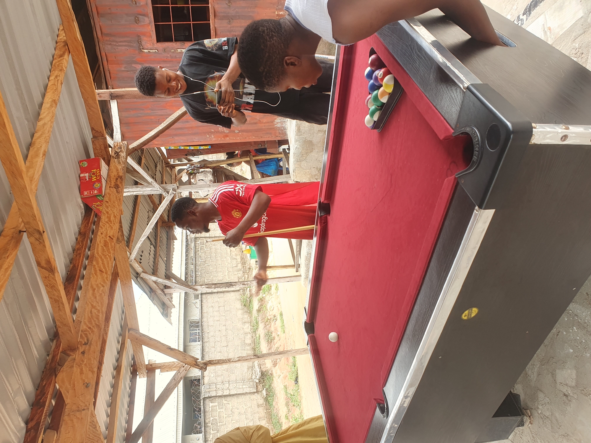 The image shows a group of young people gathered around a red pool table. The scene appears to be an outdoor recreational area, possibly a backyard or a local spot for games, under a rustic wooden and corrugated metal roof structure. The setting is consistent with a location in Lokoja, Nigeria, which is known for its vibrant community life and informal gathering spaces.
In the foreground, a dark-skinned young woman in a white top is intensely focused on the pool table, leaning forward as if about to play. To her right, a triangular rack of pool balls is neatly arranged. A single white cue ball rests in the middle of the table.
Behind the table, two young men are interacting. One, wearing a red Manchester United jersey, is holding a pool cue and appears to be in the process of playing or explaining a shot. He is looking towards the table. Another young man, wearing a black t-shirt and earphones, is holding a smartphone and smiling, looking down at the device.
The environment features a brick wall and barbed wire fence in the background, suggesting a semi-urban or residential area. The overall impression is one of casual leisure and social interaction.