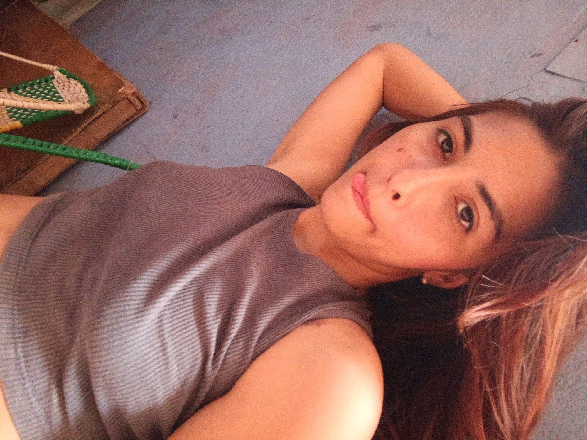A person is lying down, facing the camera. The person has brown hair and is wearing a gray tank top. The person's right arm is raised above their head. The background is a textured, light gray surface. To the left is a woven hand fan on a wooden surface. The location is unknown.