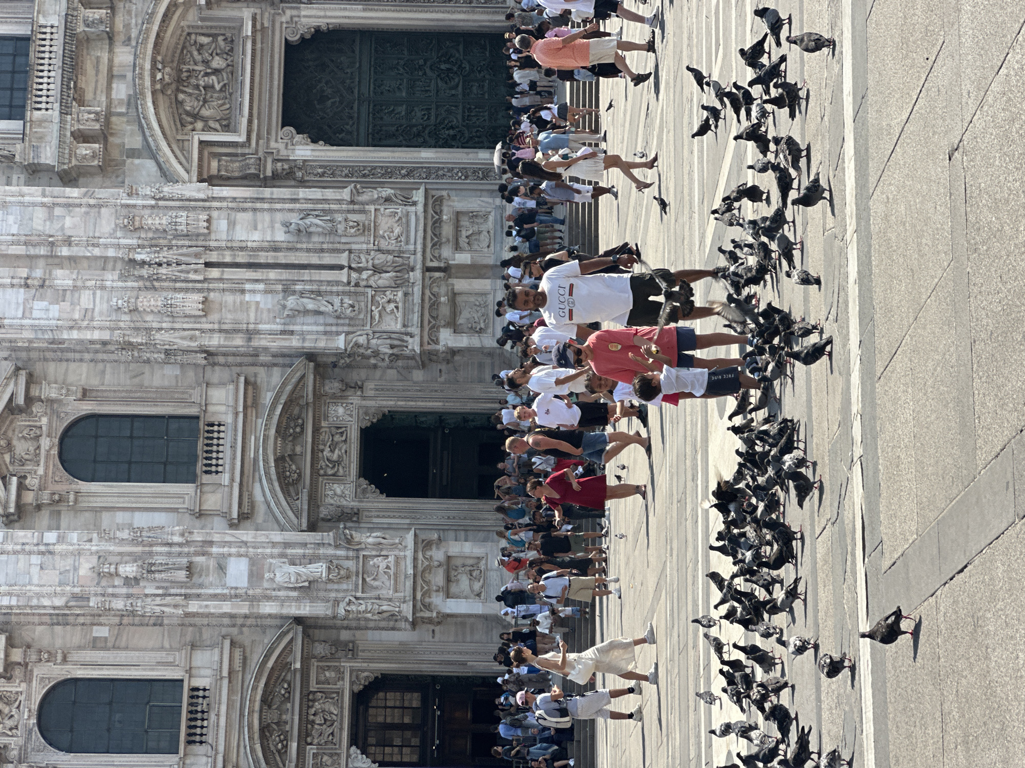 This is a medium shot, taken outdoors, of the exterior of the Milan Cathedral in Milan, Italy. The cathedral is a large Gothic structure made of white marble with intricate carvings and large arched doorways. In the foreground, a large number of pigeons are gathered on the paved plaza. Several people are walking around and interacting with each other. A man in a white t-shirt with "GUCCI" printed on it is walking towards the camera. A woman in a red dress is talking to a man in a black t-shirt and denim shorts. A young boy in a red t-shirt and a man in a white t-shirt and shorts are also visible. The sun is shining, casting shadows on the ground.
