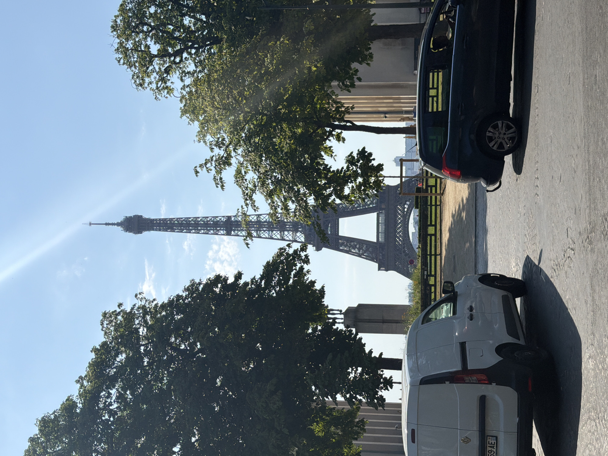 The image shows a view of the Eiffel Tower in Paris, France, on a sunny day. The tower is in the background, framed by trees on either side. In the foreground, two cars are parked on a street. A white van is on the left, and a dark-colored SUV is on the right. The street appears to be paved, and there is a shadow cast by the van. The sky is clear and blue, with a bright sun creating lens flare. The overall scene is one of a typical Parisian day, with iconic landmarks and everyday life coexisting.