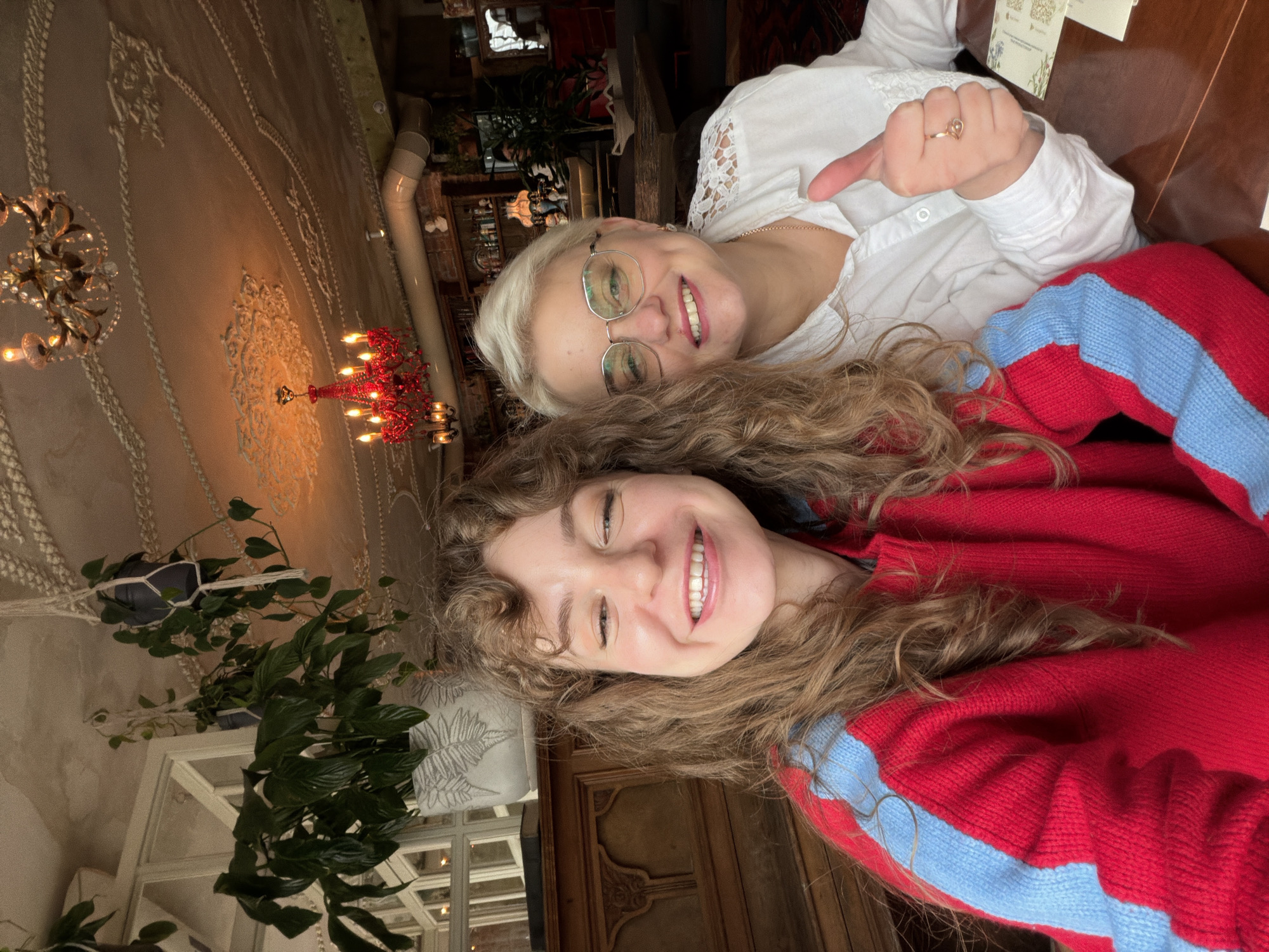 An image, oriented horizontally, captures two women smiling in what appears to be an ornate indoor setting, possibly a restaurant or cafe in Ryazan, Russia.
In the foreground, a younger woman with long, curly brown hair is visible from the chest up. She wears a red knitted sweater with prominent light blue stripes running across it. Her head is tilted slightly, and she has a wide, joyful smile, looking directly towards the camera.
Behind her and slightly to the right, an older woman with short, light blonde or white hair and glasses smiles broadly, also looking at the camera. She wears a white collared shirt with lace detailing on the shoulders and a thin gold chain around her neck. Her right hand is raised, giving a thumbs-up gesture, and a small, delicate ring with a heart-shaped design is visible on her finger.
The background reveals a richly decorated interior. The ceiling features intricate, raised patterns in a light, possibly beige or cream, color. Several chandeliers hang from the ceiling: one ornate golden or crystal fixture with multiple bulbs and another distinct red chandelier. Lush green plants in macrame hangers and on shelving are visible, adding to the warm ambiance. Dark wooden paneling or furniture is also seen towards the lower right. The overall impression is one of a cozy yet elegant establishment. Both women appear happy and relaxed, suggesting they are enjoying their time together. No legible text is visible in the image.