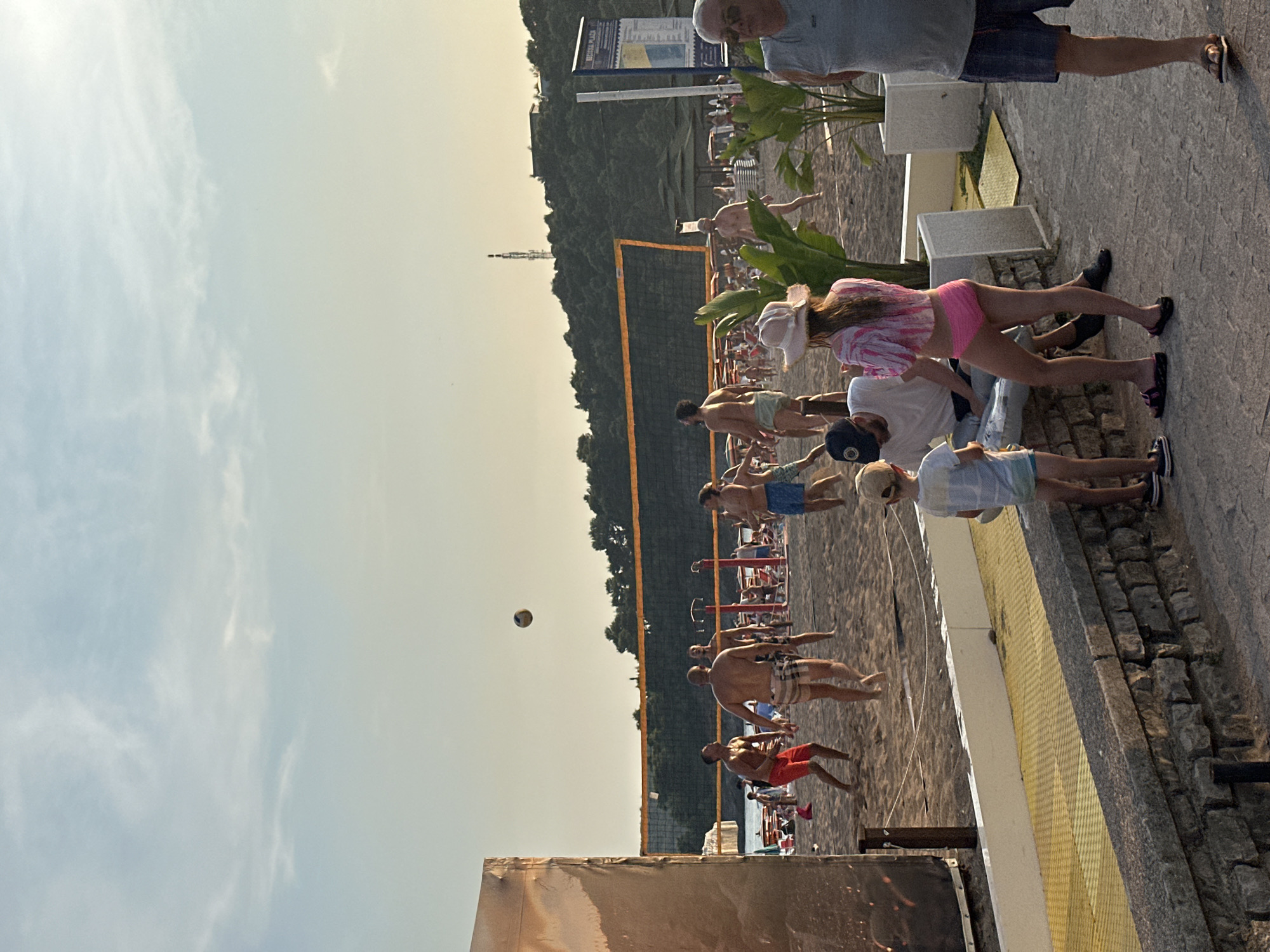 The image captures a lively scene on a sandy beach, likely in Boreti, Montenegro, with a volleyball game in progress under a clear sky. Several men are actively playing volleyball near a net, some in mid-air as they jump to hit the ball. Spectators are present on the beach, with people sitting on lounge chairs and others standing around. A young child is standing on a paved walkway in the foreground, looking towards the game. Further in the background, trees and hills form a scenic backdrop. The overall atmosphere suggests a recreational and social gathering on a sunny day.