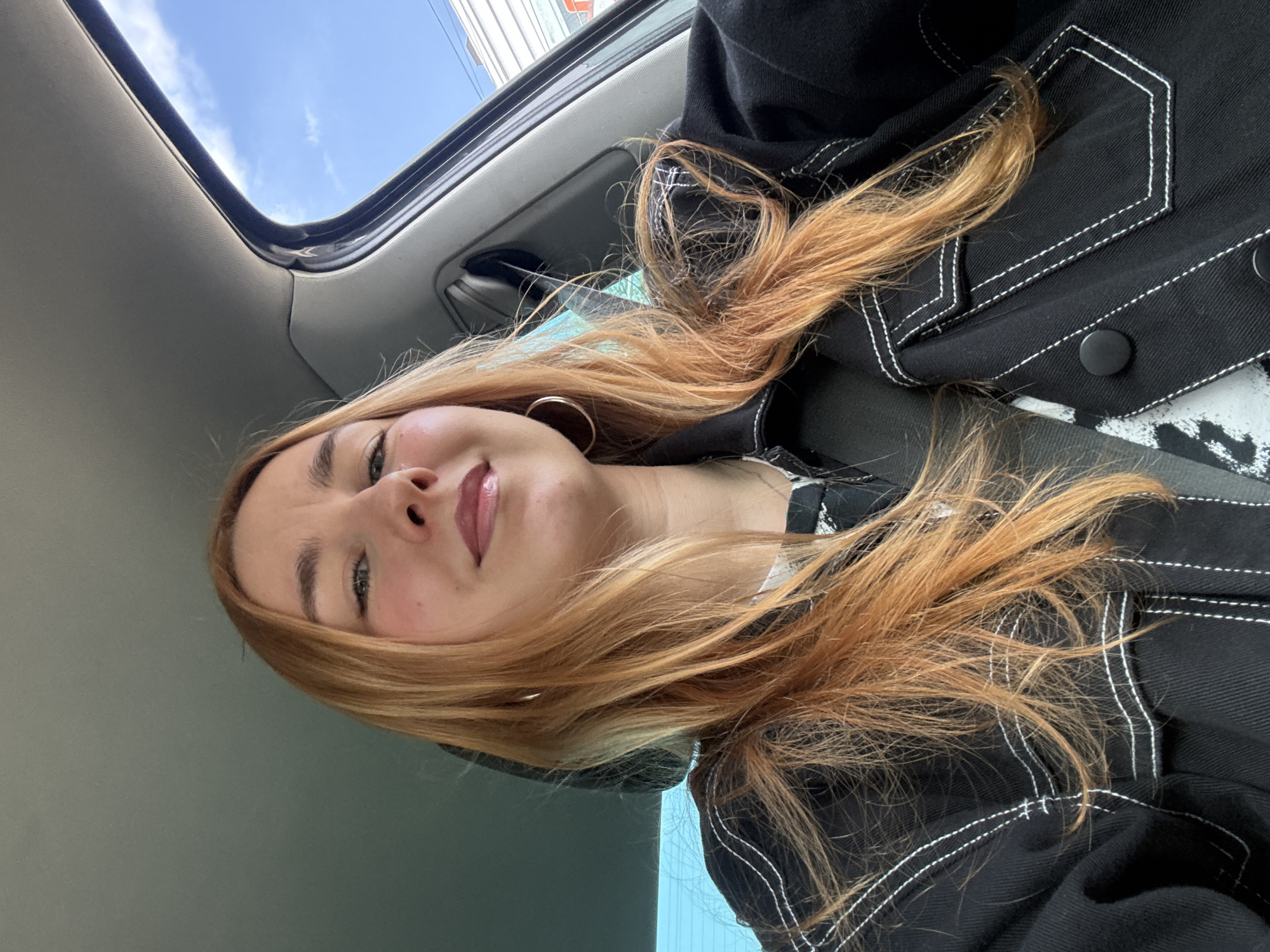 A young woman is pictured inside a car, looking towards the camera. She has light skin, green eyes, and reddish-blonde hair. She is wearing hoop earrings and a black denim jacket with white stitching. A car sunroof is visible, showing a view of a blue sky with some clouds. The location is not possible to discern from the image. The city of Minsk, Belarus is not possible to identify.