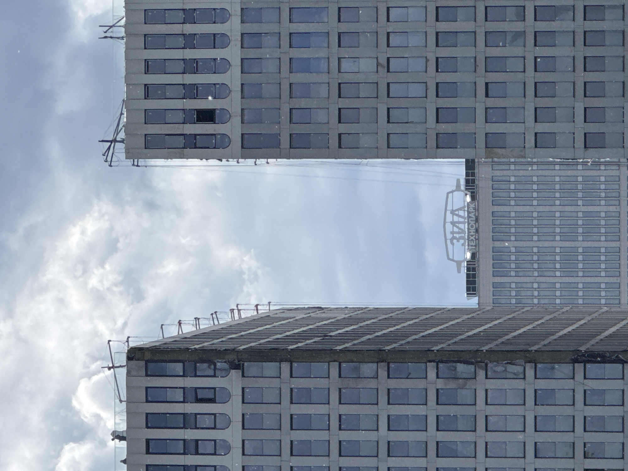 Two tall, grey high-rise buildings stand prominently against a cloudy sky, captured from a low-angle perspective looking upwards between them. The buildings feature a repetitive grid of rectangular windows, some of which have distinctive rounded tops, particularly noticeable on the left building. The facades appear to be made of concrete or a similar stone material.
On the upper edges of both buildings, there are visible structures resembling scaffolding or construction equipment, suggesting ongoing construction or maintenance work.
A significant detail is the visible text on the side of the right building. In Cyrillic script, the word "ТЕХНОПАРК" (TECHNOPARK) is clearly readable, with "3Д" (3D) partially visible above it.
The sky is bright but filled with scattered white and grey clouds, indicating daytime conditions. There are no people visible in the image. The scene conveys an urban environment, and with the provided context, the location is Moscow, Russia.