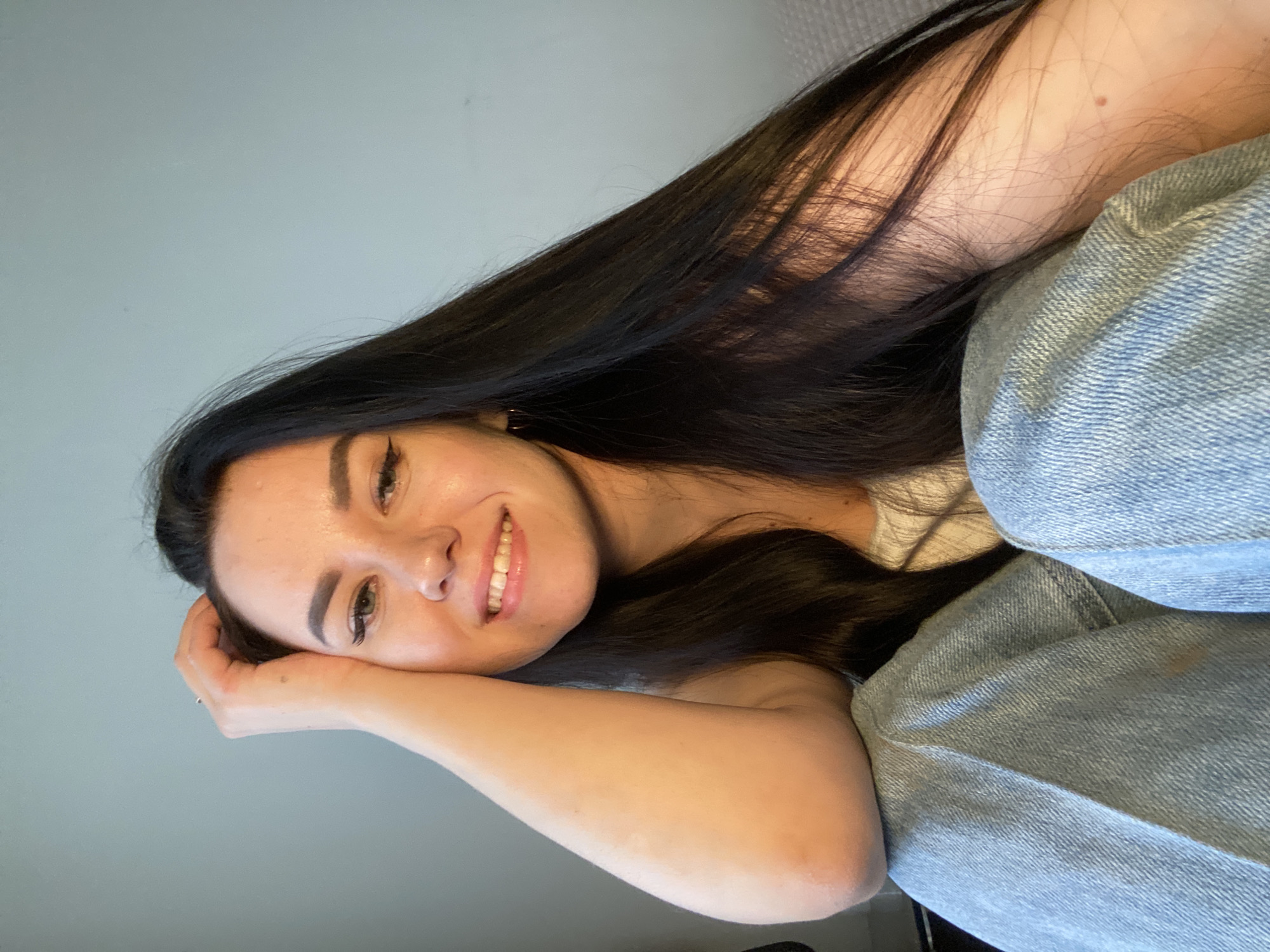 The image shows a young woman lying on her side, looking directly at the camera with a gentle, relaxed smile. She has long, dark hair that frames her face and extends over her shoulder. Her head is supported by her right arm, which is bent at the elbow. She is wearing light blue denim jeans, with their textured fabric visible in the lower right of the frame. The background consists of a plain, light greyish-blue wall, indicating an indoor setting, possibly on a sofa or bed.
Her expression conveys a sense of ease and happiness. She has well-defined eyebrows, visible eyeliner or mascara, and natural-looking lip makeup. A small mole is noticeable on her upper right arm or shoulder. The lighting is soft and even, suggesting either natural daylight from an unseen window or diffused indoor lighting. There are no other significant objects or visible text in the image. The specified context for this media file is Chaikovsky, Russia.