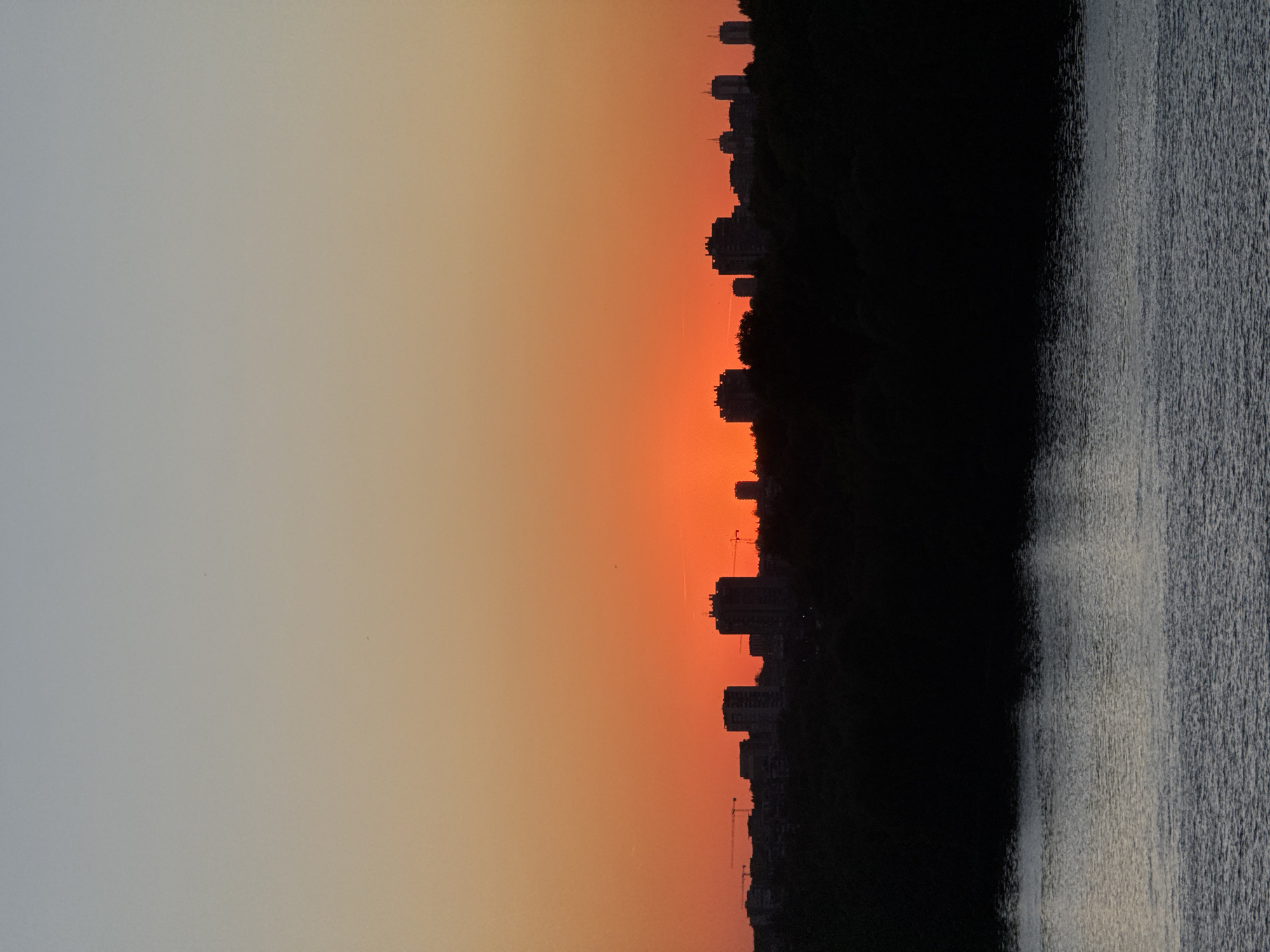 The image captures a cityscape at sunset. The left side of the frame displays a gradient sky, transitioning from pale gray to orange. A line of buildings and trees is silhouetted against the orange sky. A dark, undefined mass occupies the right side of the frame, textured like water. Two cranes are visible among the building outlines. The location is likely Belgrade, Serbia.