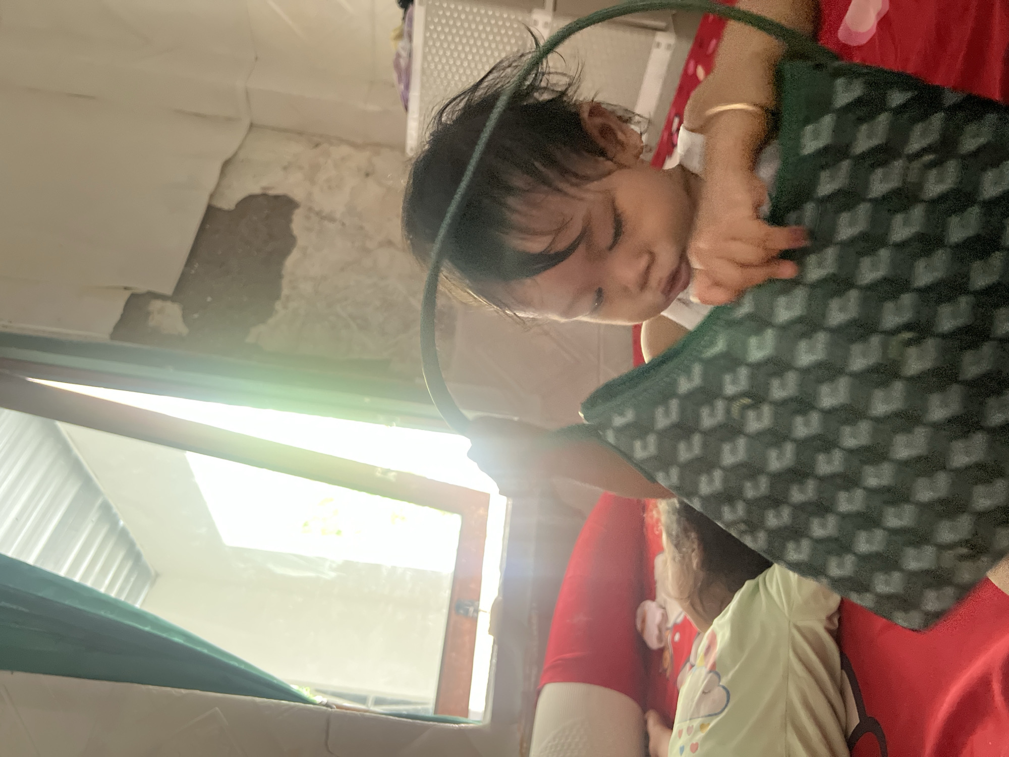 A young child with dark hair is shown looking into a dark green bag with a geometric pattern. The child is wearing a white top and a gold bracelet on their left wrist. The background includes a window with a bright, daytime view outside, a wall with peeling paint, and a white lattice object. Another child is lying down on a red surface under the bag. The photo appears to be taken inside a residence. The location is Kecamatan Pati, Indonesia.
