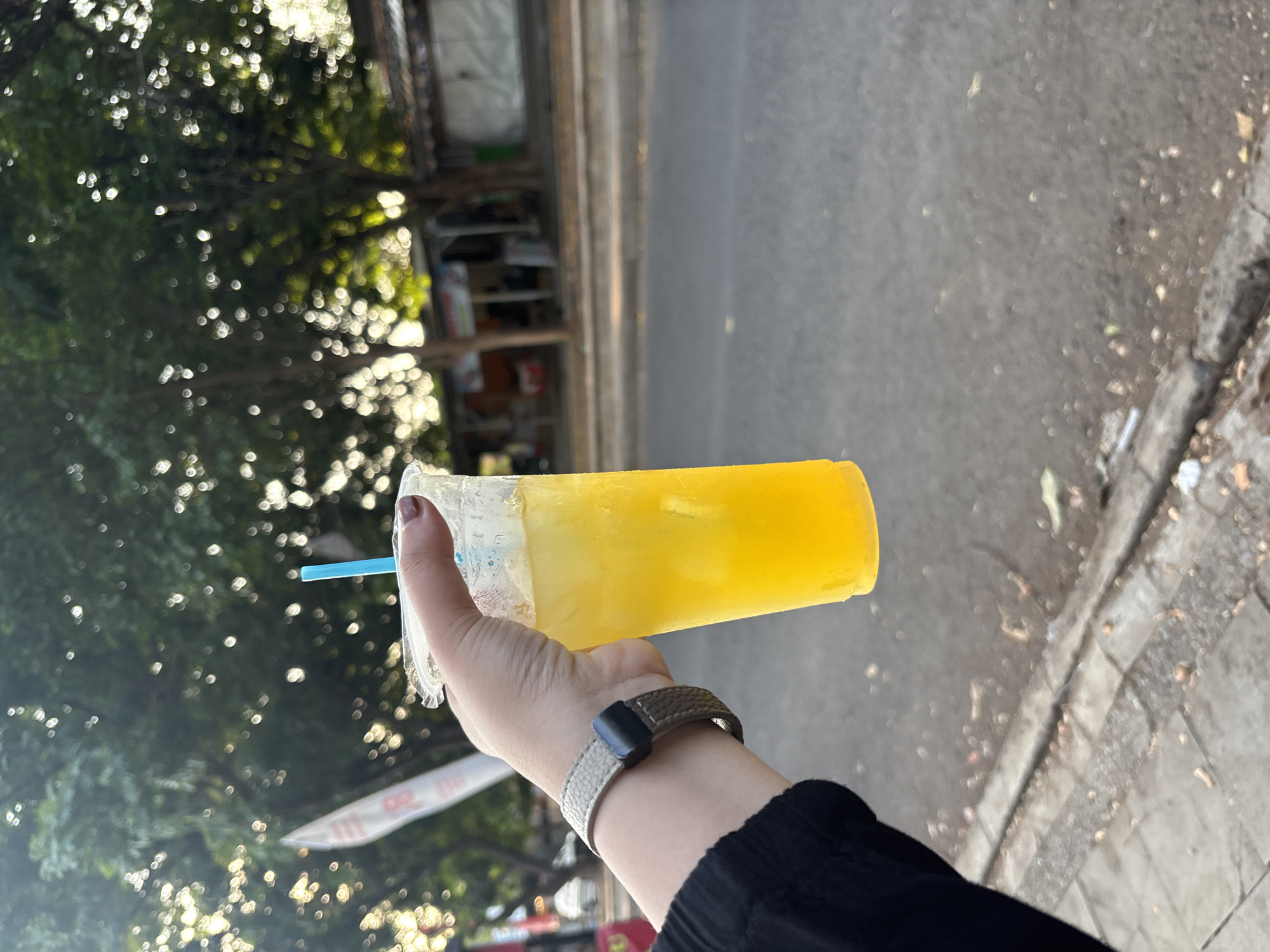 An individual's hand, adorned with a watch on the wrist and dark nail polish, holds a clear plastic cup filled with a bright yellow, iced beverage. A blue straw is visible in the drink. The scene appears to be outdoors, with a blurred background of green trees and a street. The setting suggests a location in Cirebon, Indonesia, possibly a roadside or pedestrian area, with a glimpse of shops or stalls in the distance. The lighting indicates it is daytime, with sunlight filtering through the trees. The overall impression is casual and candid.