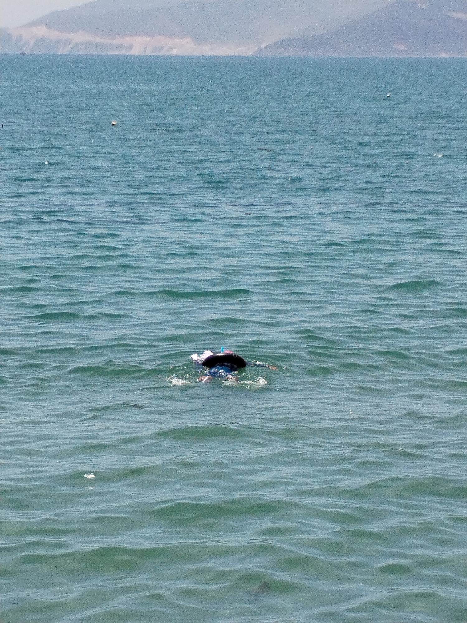 The image shows a person snorkeling in the sea. The individual is wearing a dark cap and snorkel. They are surrounded by calm, rippling turquoise water. In the distance, the coastline of Bac Nha Trang Ward, Vietnam, is visible as a faded mountain range. The weather appears clear.