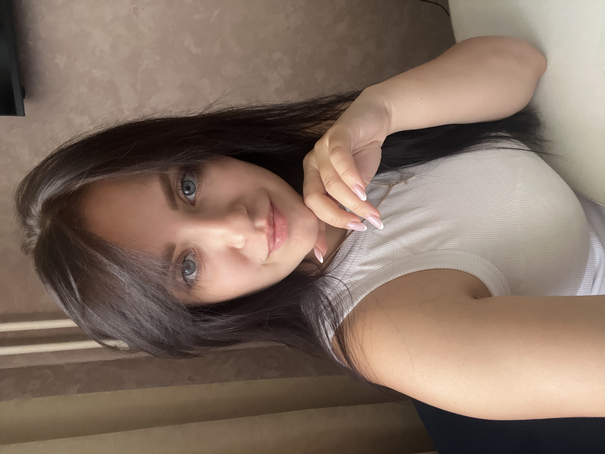 In the foreground, a young woman with dark hair and blue eyes is captured in a close-up selfie. She is wearing a white ribbed tank top and resting her chin on her hand. Her nails are manicured with a French tip design. A delicate gold necklace with a cross pendant is visible. The background consists of a textured wall and vertical striped curtains. The image appears to be taken indoors, likely in a residential setting. The lighting suggests it is daytime. The location of Марковский, Russia is not explicitly depicted or inferable from the visual elements within the image.