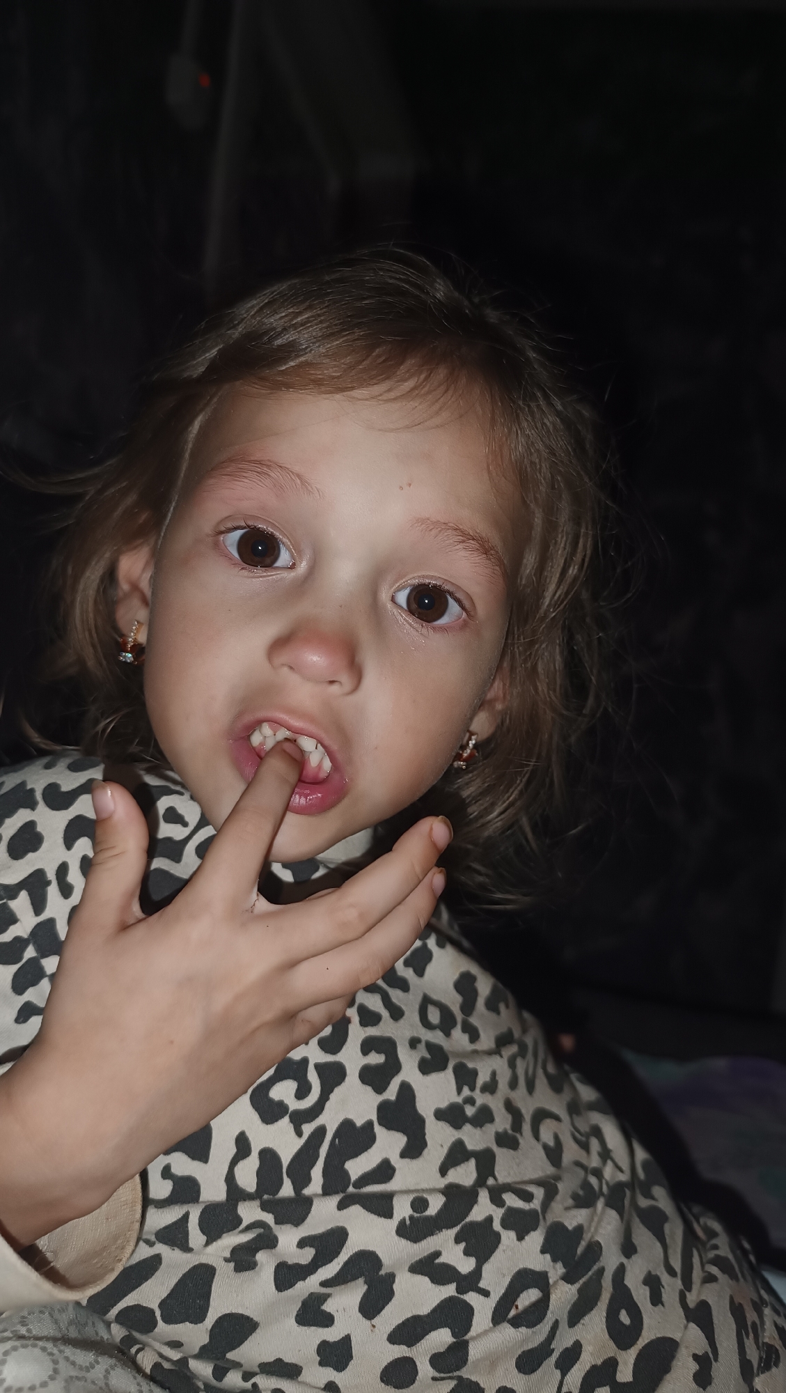 A close-up, head-and-shoulders shot shows a young girl with light brown, wavy hair, her mouth open and a finger touching her teeth. She is wearing a light-colored top with a black leopard print. Small, dark earrings with red accents are visible in her ears. The background is dark and out of focus. There is no indication of the city of Karsha, Russia, in the image.