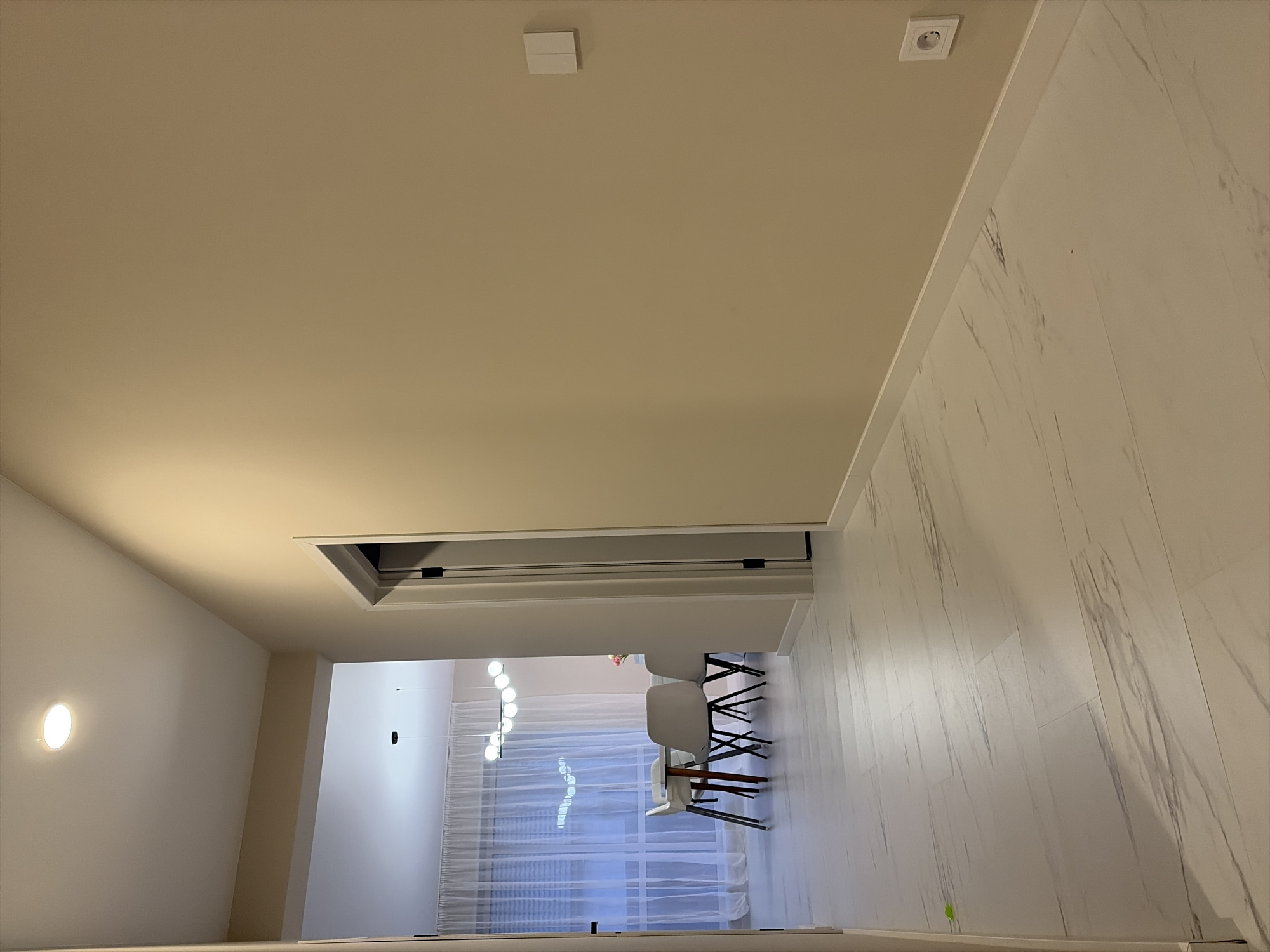 The image captures an interior view of a modern living space, primarily focusing on a light-colored hallway that transitions into an open-plan room. The perspective is from within the hallway, looking towards the deeper part of the residence.
The walls and ceiling of the hallway are painted in a uniform, light beige or cream color. On the ceiling, a small, square white fixture, possibly a smoke detector or sensor, is visible towards the left. Further to the right, a larger, square access panel or vent is recessed into the ceiling. An electrical outlet with a white cover is also mounted on the ceiling near the right edge. A circular, flush-mounted wall light illuminates the left side of the hallway.
The hallway opens into an adjacent room, likely a dining area or living space. In this room, part of a table with at least two modern chairs is visible. The chairs feature white, molded seats and black, slender legs. Above the table area, a contemporary pendant light fixture with multiple spherical bulbs hangs from the ceiling. The back wall of this room features a large window or glass door covered by translucent white sheer curtains, filtering natural light into the space.
The floor throughout the visible area is composed of light-colored, rectangular tiles or panels featuring subtle grey marbling or streaking patterns, extending from the hallway into the other room. The overall aesthetic is clean, minimalist, and brightly lit by both artificial fixtures and diffused natural light. No people are present, and no specific activity is taking place. There is no visible text in the image.