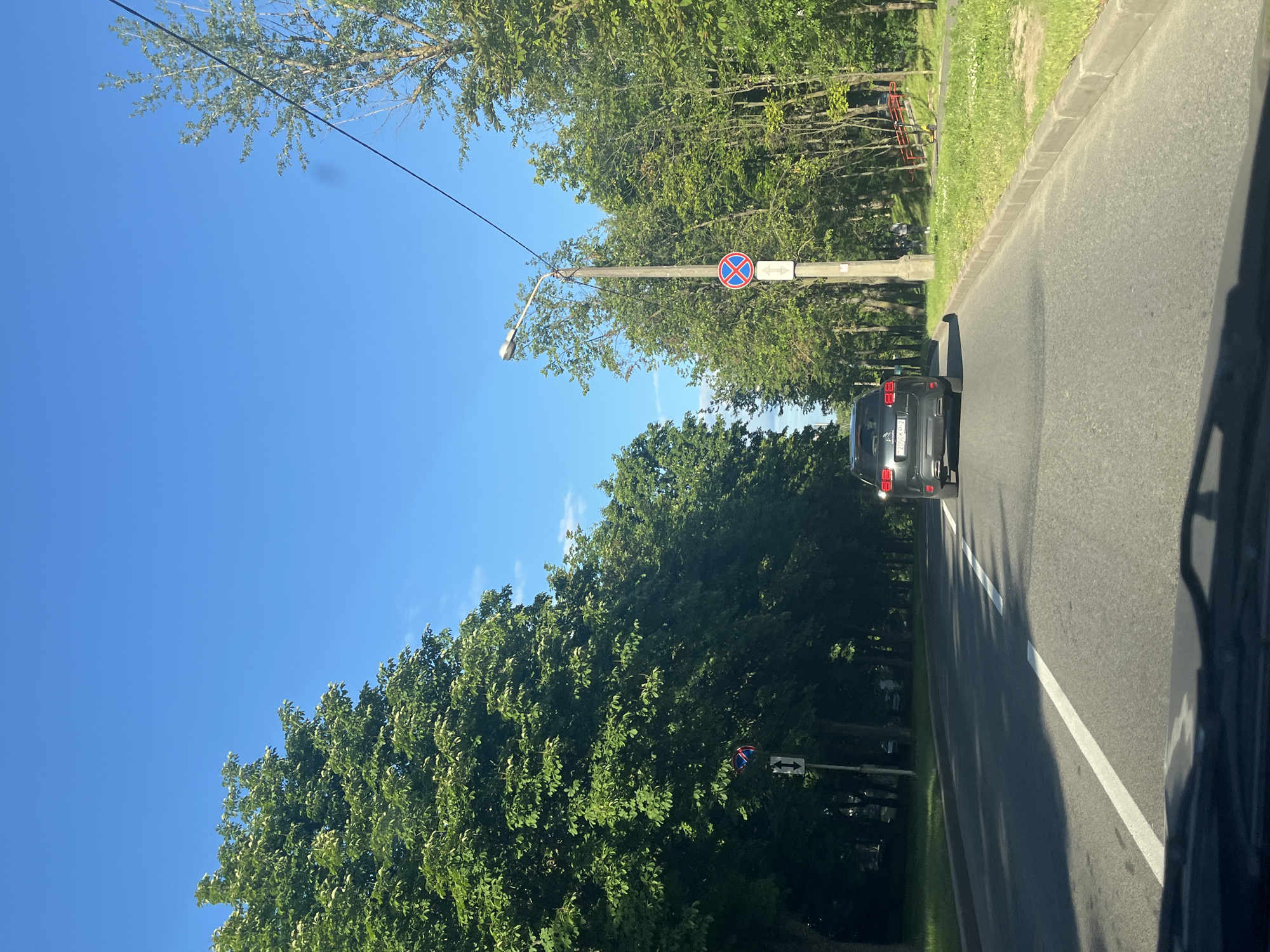 A dark grey SUV with license plate 2050 KP-2 is driving down a road lined with mature trees. The road has a white dividing line and is bordered by a grassy area. A lamppost with a "no stopping" sign and an arrow pointing down is visible to the right of the SUV. The sky is clear and blue. The scene appears to be a typical sunny day in a city park or residential area. The location is Minsk, Belarus.
