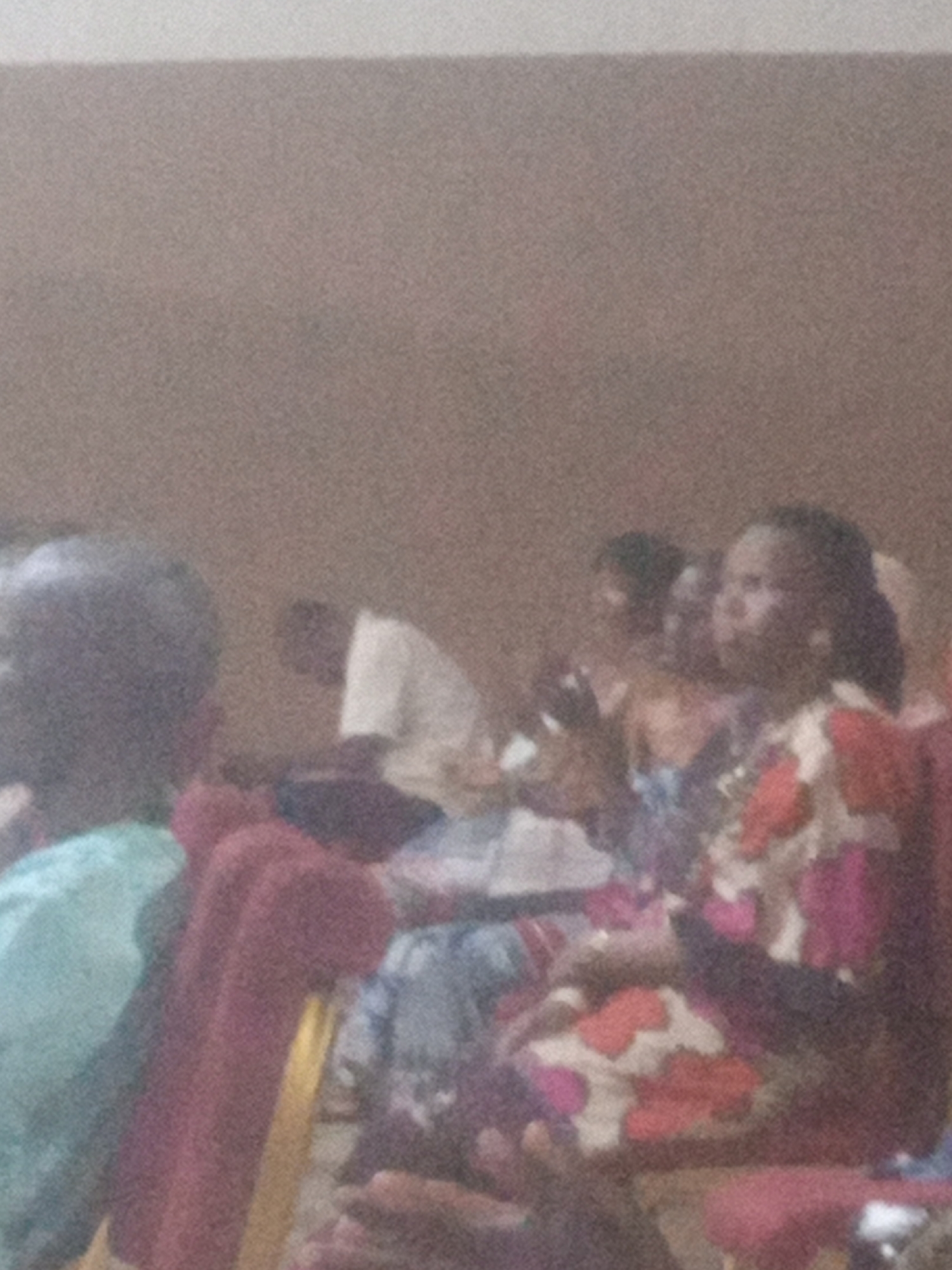 The image shows a gathering of people in what appears to be an indoor setting. The lighting is dim, and the image quality is poor, making it difficult to discern fine details. In the foreground, on the left, is the back of a person's head and shoulder, wearing a light blue garment. To the right of this person, there are several individuals seated. One person in the center is wearing a white shirt. Further to the right, a woman in the foreground is wearing a colorful patterned dress with abstract shapes in pink, white, and orange. Her face is partially visible, and she appears to be looking to her left. Other people are present in the background, but they are out of focus and their features are indistinct. The overall impression is of a community gathering or a meeting, possibly in a hall or a similar venue. The background appears to be a plain, textured wall. The location of Rukuba Road, Nigeria, is mentioned as context but is not visually identifiable from the image itself.