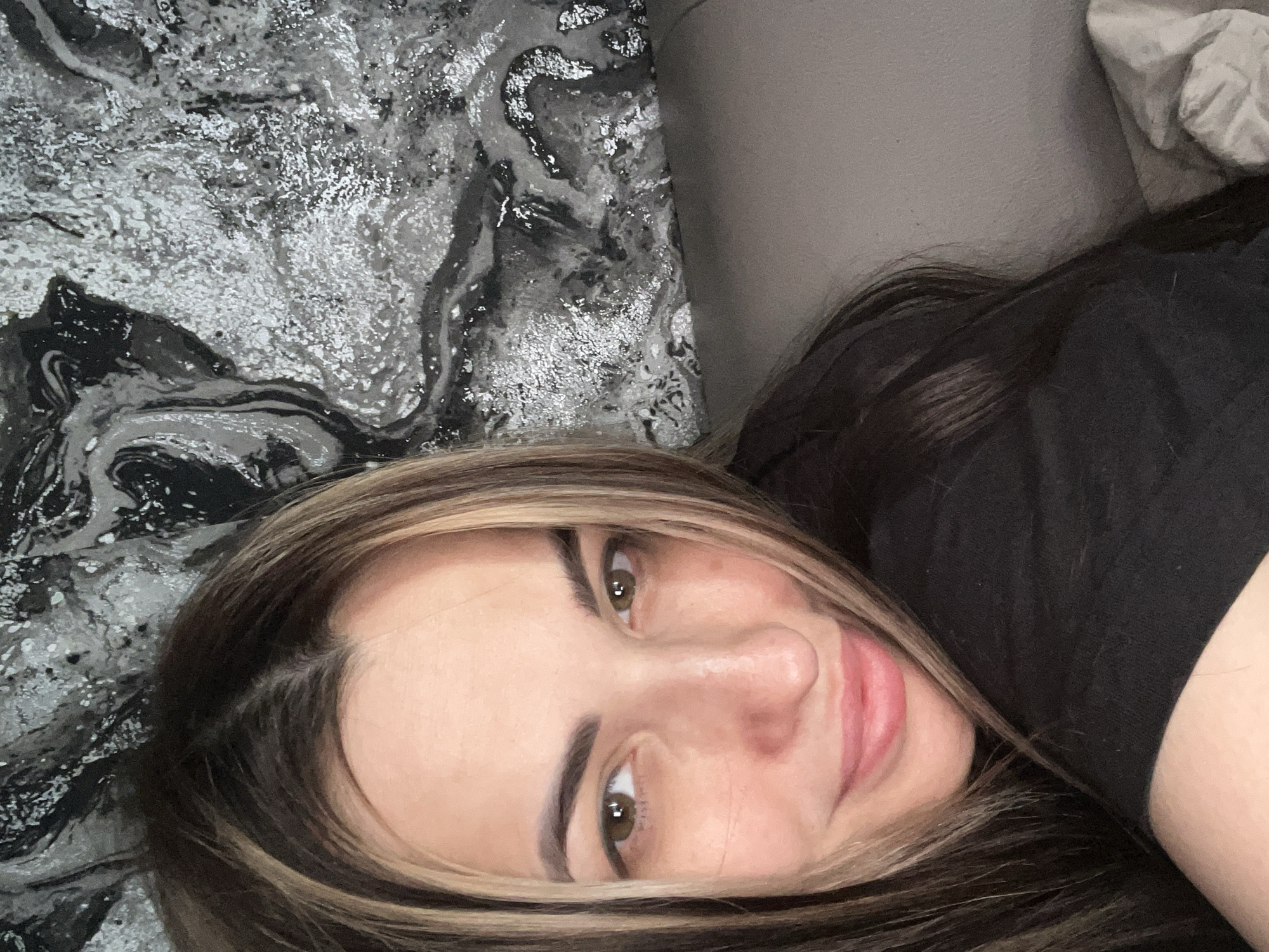 A close-up, angled view captures a person with fair skin, brown hair highlighted with blonde streaks, lying down on what appears to be a bed or couch. The person is wearing a black shirt. The background includes a gray, marble-patterned surface and a plain gray surface, potentially a wall or another part of the furniture. The location is Unknown, Unknown.