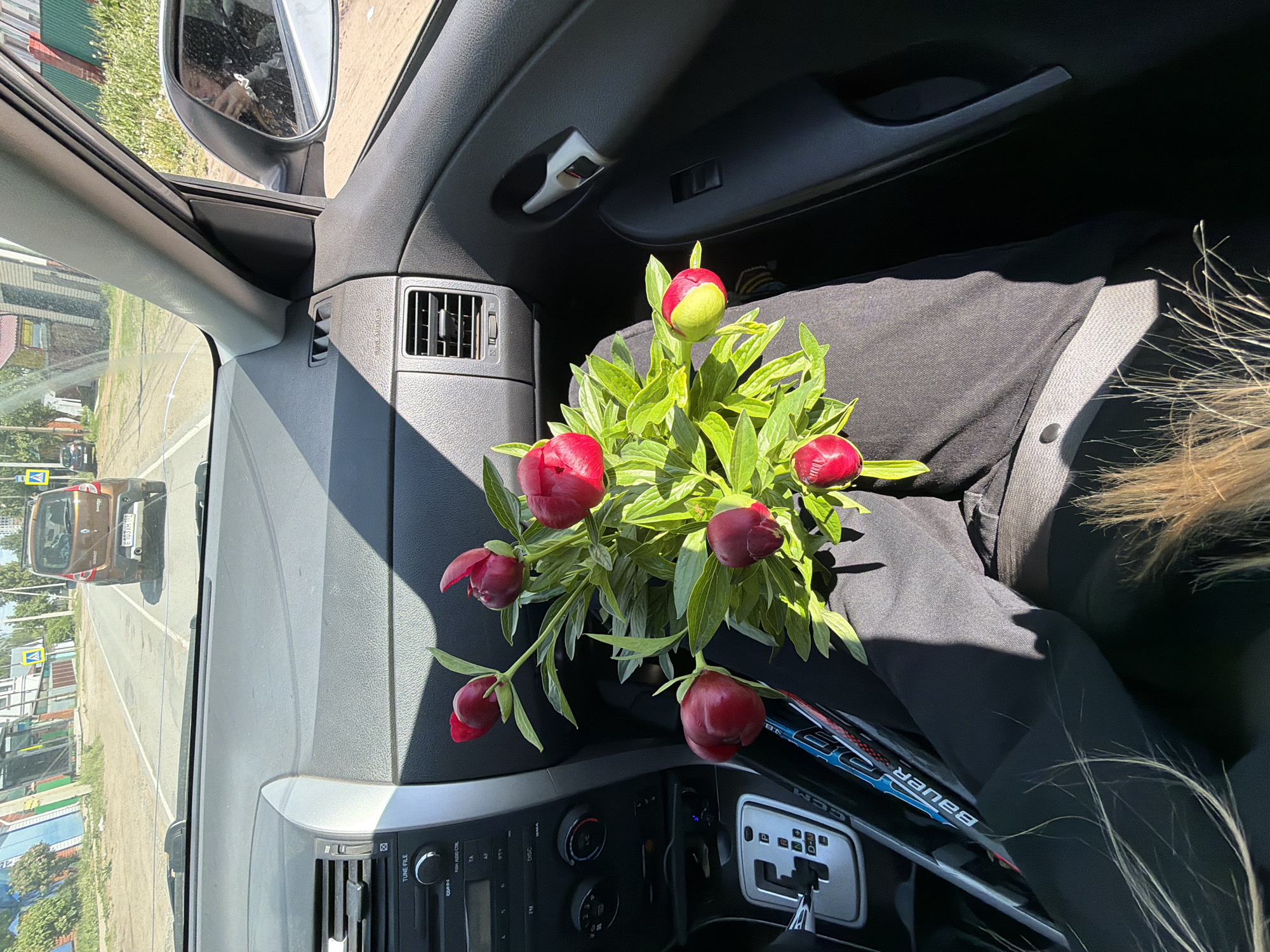 The interior of a car is shown. On the passenger seat rests a bouquet of red peonies with green foliage. Part of a woman's blonde hair is visible near the right edge of the frame, and what appears to be a hockey stick lies next to the bouquet. Through the windshield, a street scene in what appears to be the city of Chaikovsky, Russia, is visible, with a brown car on the road.