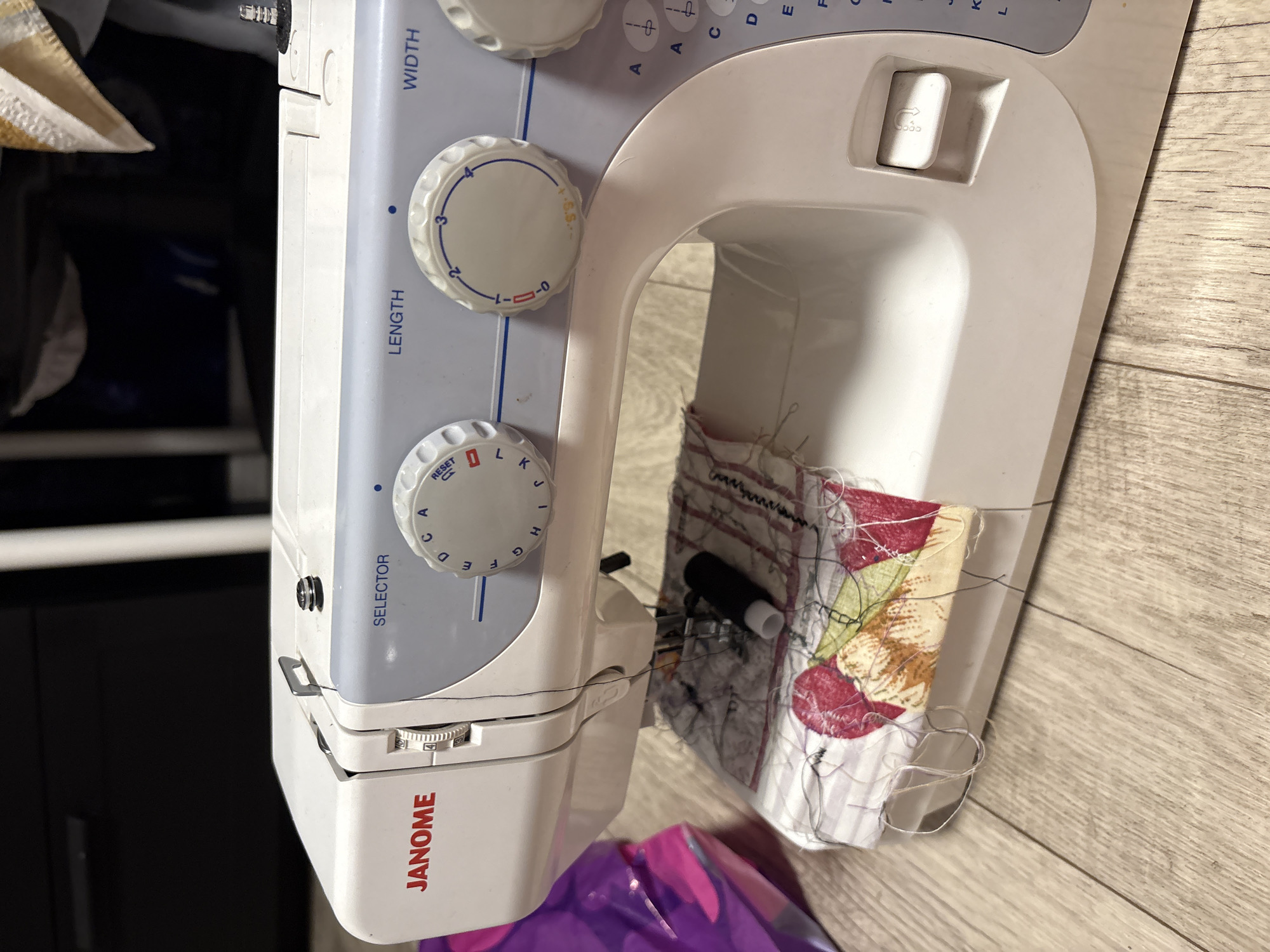 An indoor, close-up photograph displays a white and light blue Janome sewing machine positioned on a light-colored wooden surface, likely a floor or table. The machine is oriented vertically from an overhead, slightly tilted perspective. Fabric remnants, adorned with colorful patterns in red, yellow, and white, are loosely tucked into the free arm of the machine, covered in numerous black and white threads, suggesting a current or recently finished sewing project. A small black object, possibly a bobbin or marker, is also visible among the fabric. A purple plastic bag is partially visible at the bottom left, and dark furniture elements are blurred in the background on the left.
The sewing machine features several control dials and buttons. Visible text includes "JANOME" prominently displayed on the front base. Dials are labeled "SELECTOR" (with letters A through L and "RESET"), "LENGTH" (with numbers 0 through 4 and "S.S."), and "WIDTH" (with numbers -0- through 4). Faintly, the word "PRESS" is visible on a rectangular button on the top right side of the machine. The overall scene depicts a functional, hobbyist or crafting workspace. No individuals are present, and the indoor lighting provides no specific cues about the time of day or weather.